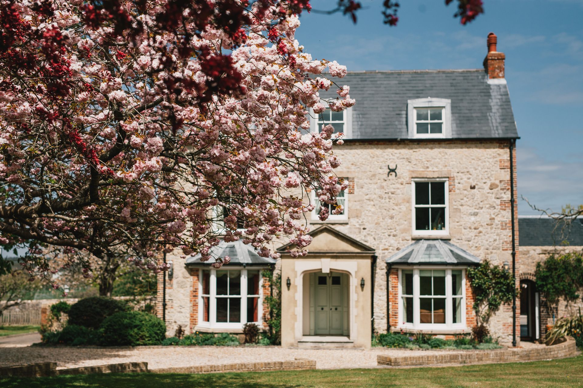 Stone house with gray roof, flanked by flowering tree, two bay windows.