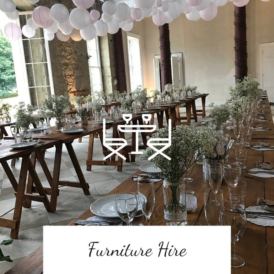 Long wooden tables set for a gathering, white decor, and paper lanterns in a large room.