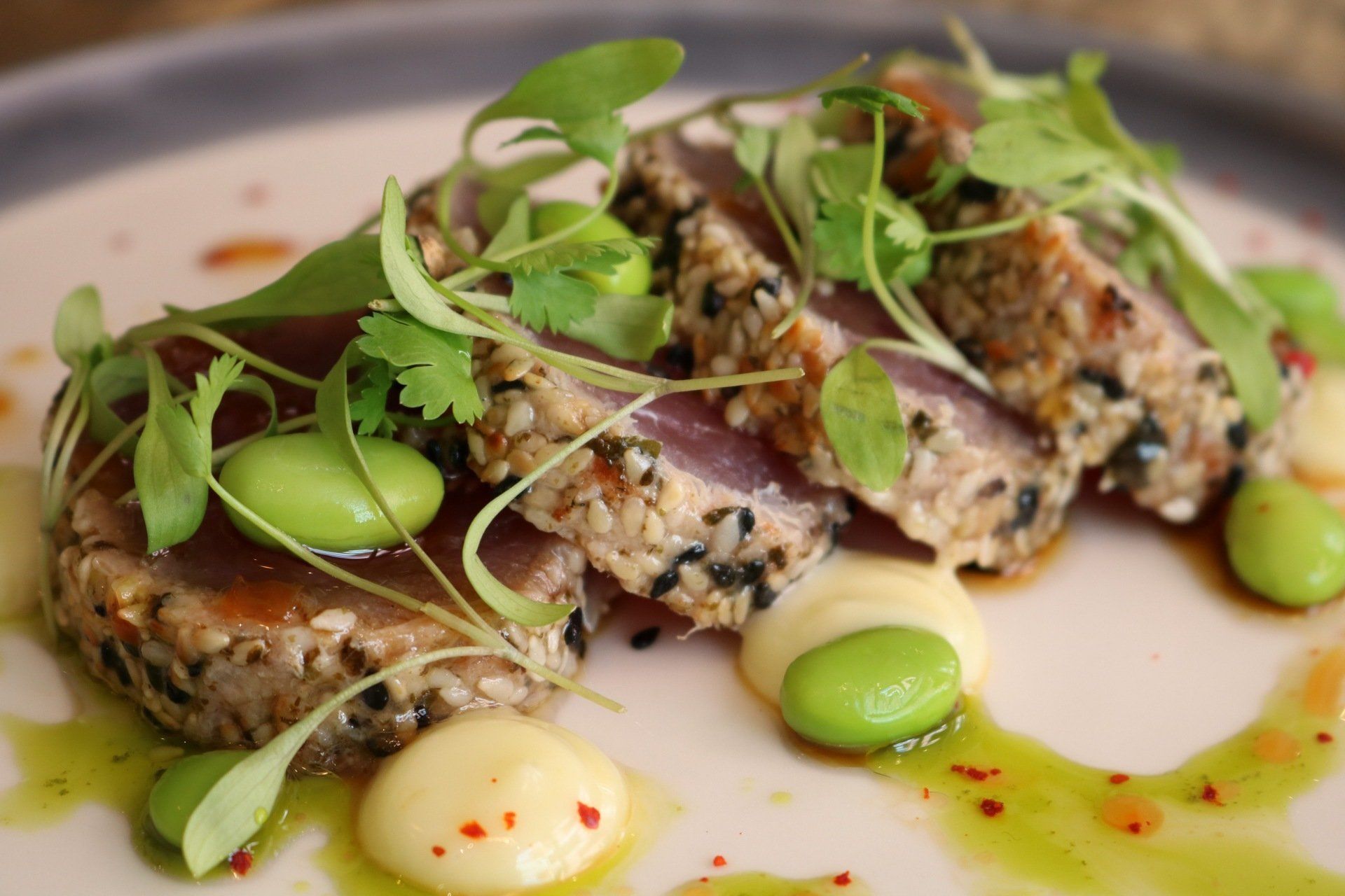 Seared tuna with sesame crust, edamame, microgreens, and sauce on a white plate.