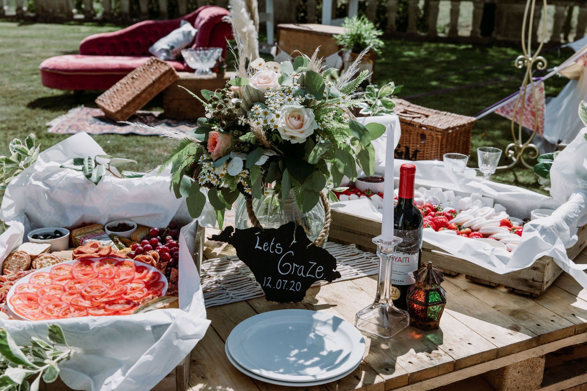 Outdoor charcuterie spread with flowers, wine, and a red chaise lounge.