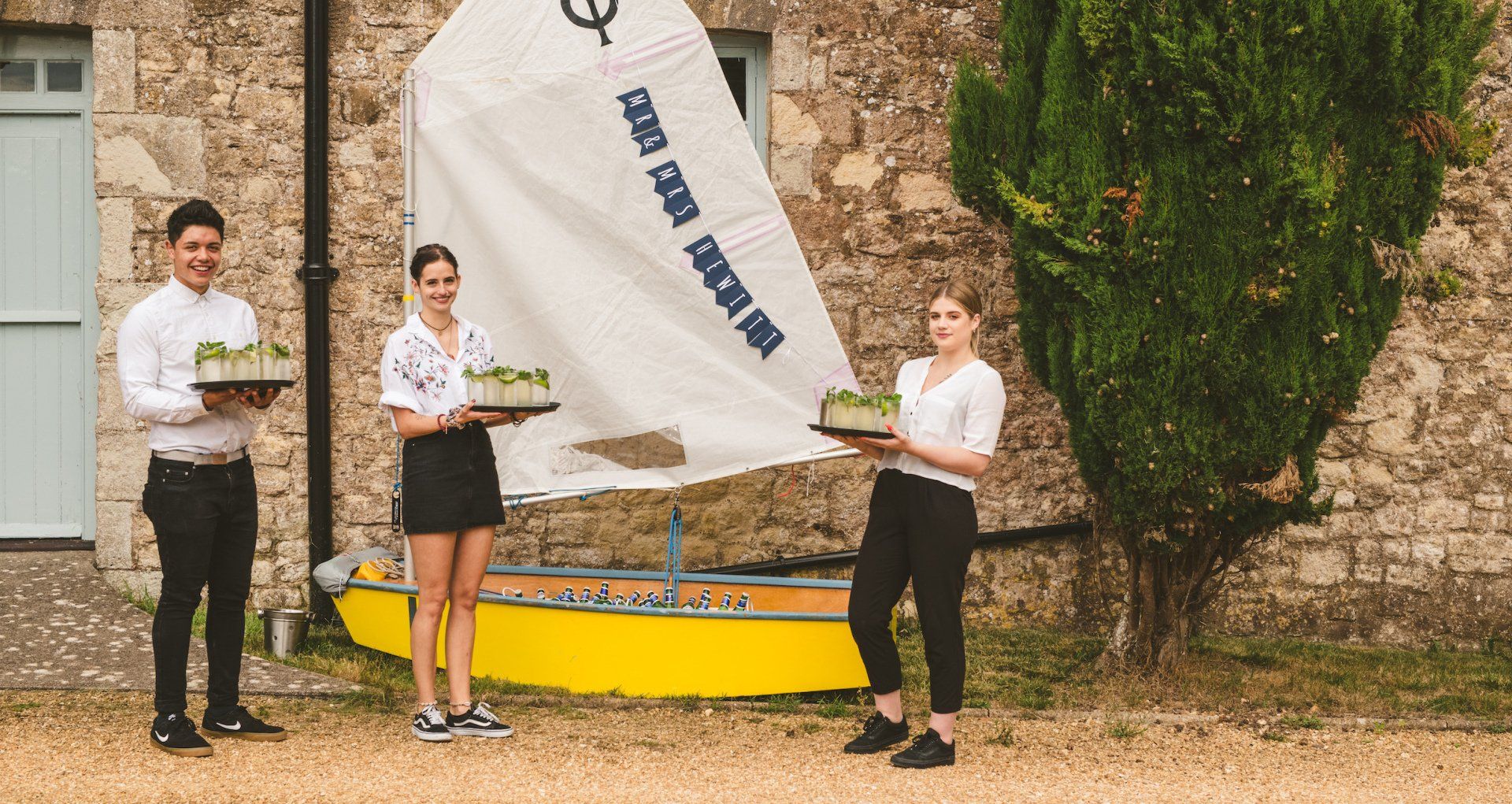 Three people hold cakes in front of a yellow sailboat, standing next to a stone building.