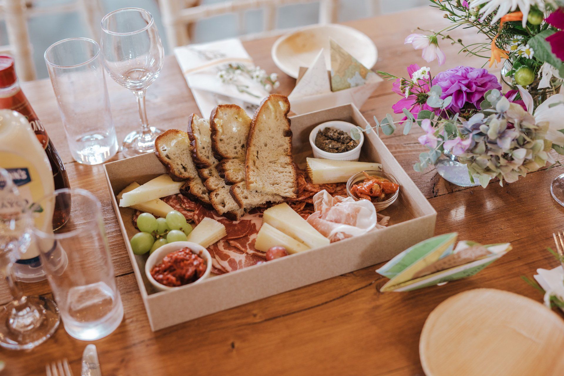 Charcuterie board with bread, meats, cheese, grapes, and dips on a wooden table, next to flowers and glasses.
