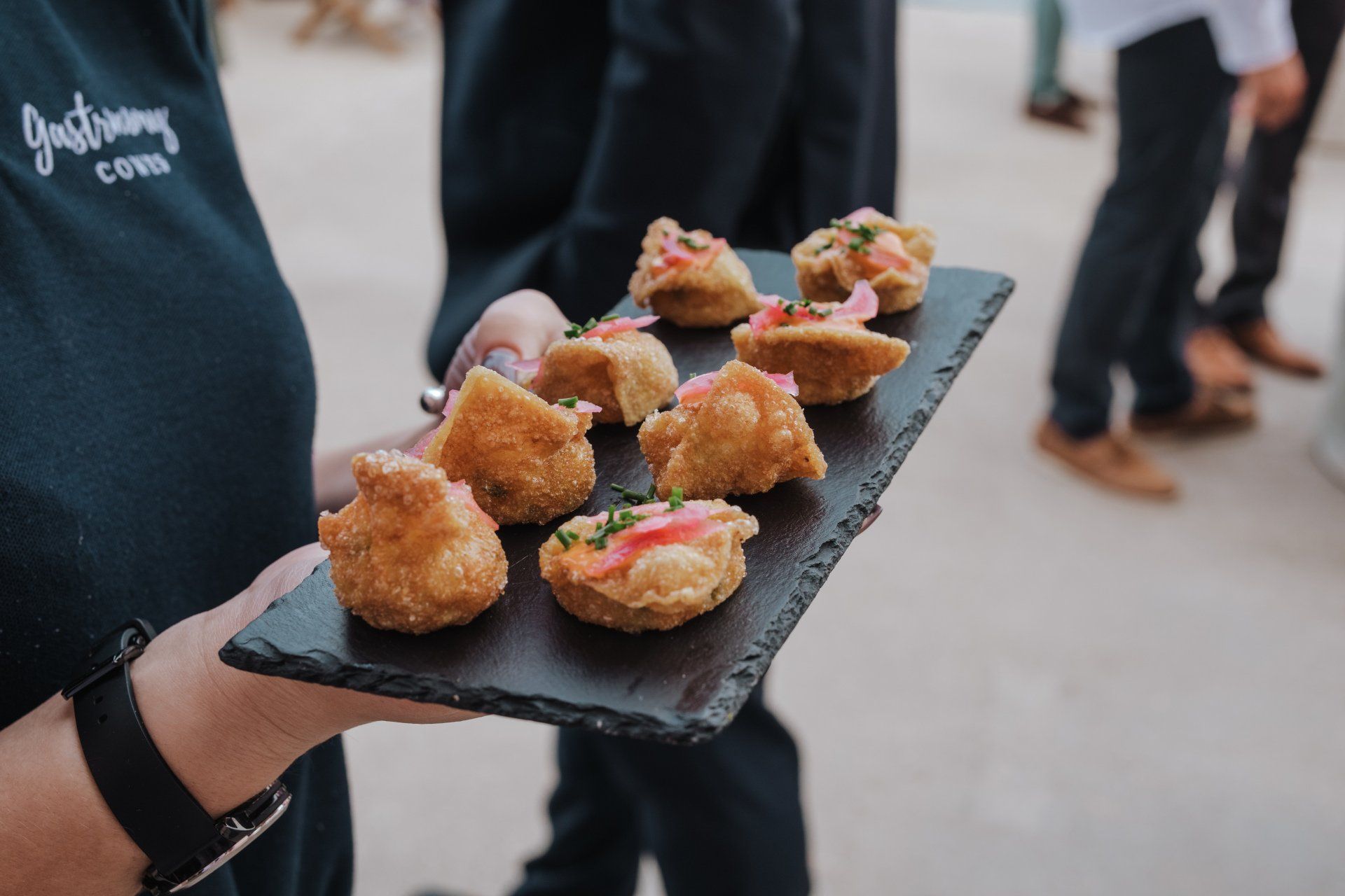 Server holding a slate tray with fried food appetizers, topped with pink garnish.
