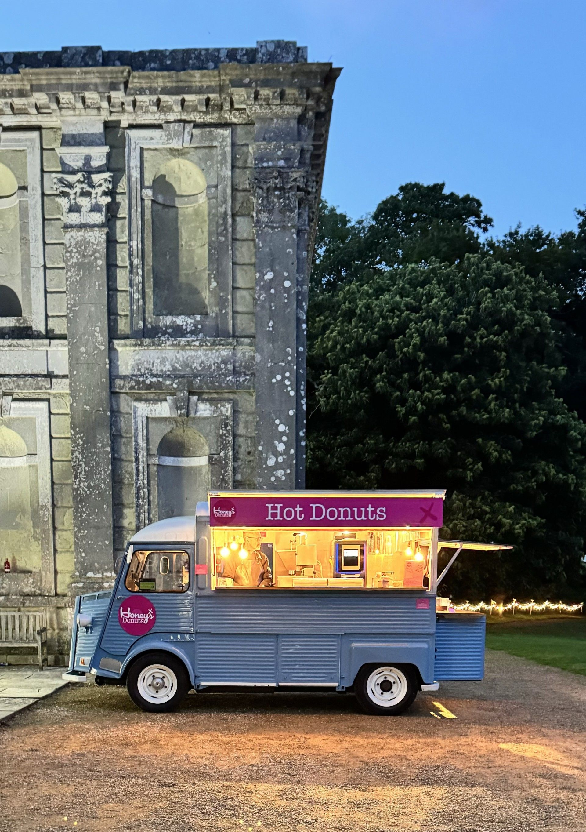 Blue food truck selling hot donuts outside a stone building at dusk.