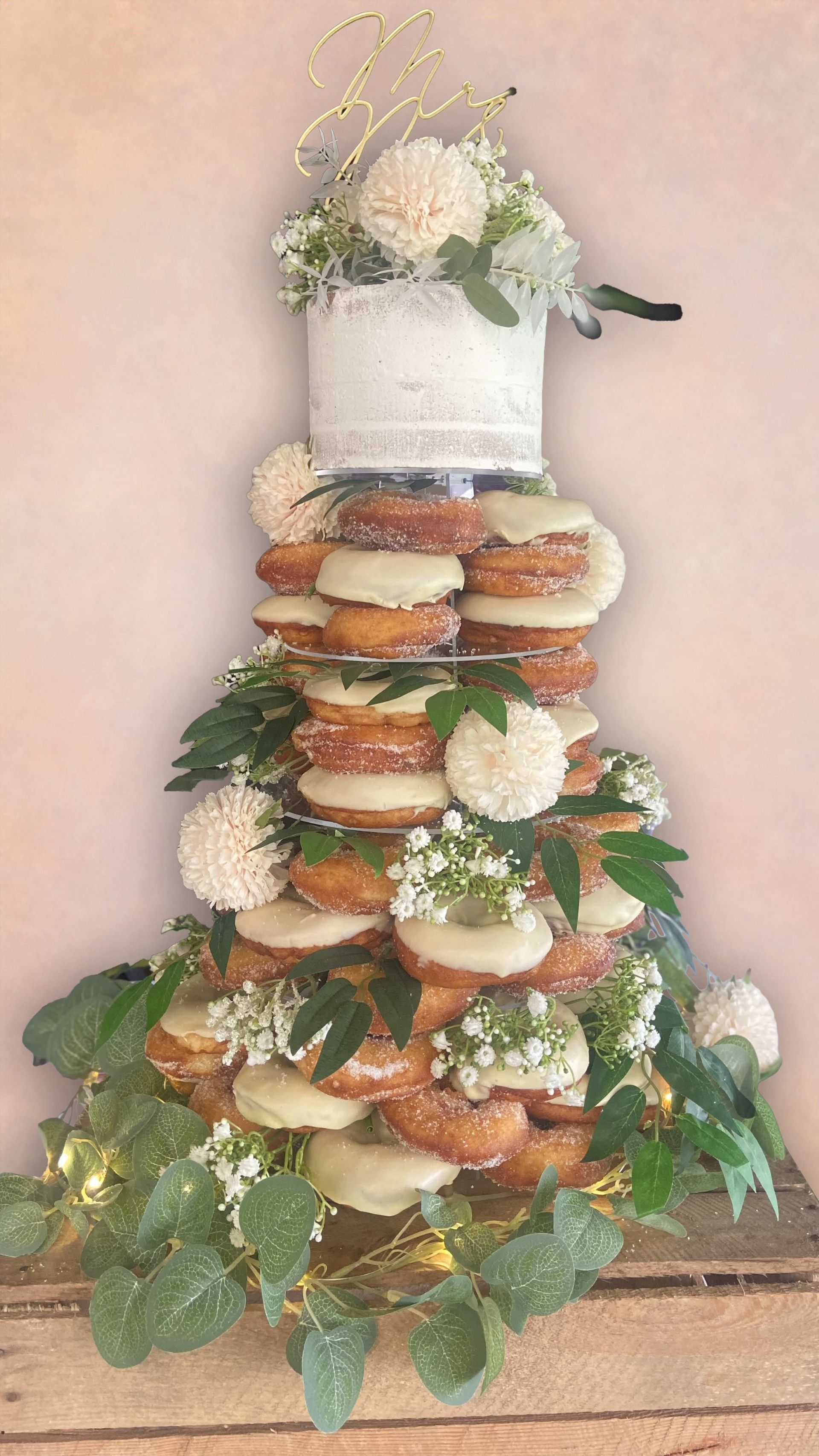 Donut tower decorated with greenery, flowers, and a small cake on top; a wedding treat.