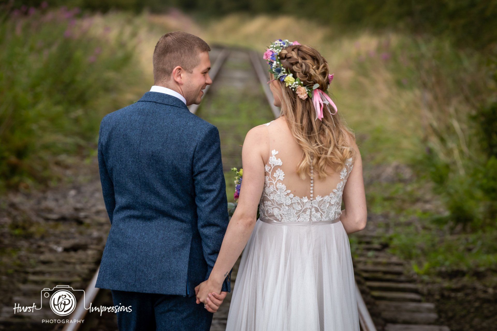 Couple holding hands, walking on railroad tracks, she wears a floral crown and wedding dress, he wears a suit.