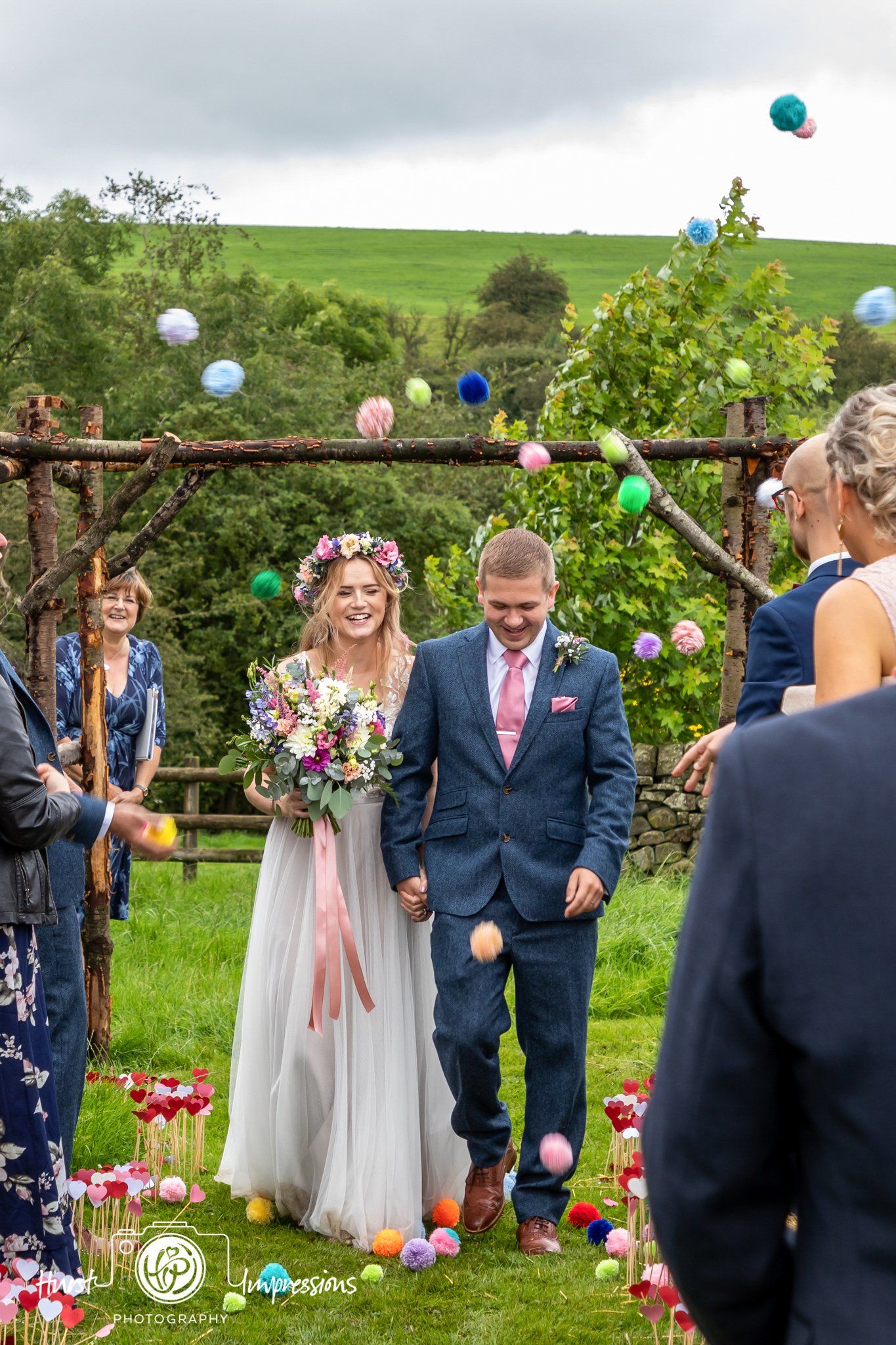 Newly married couple walks down aisle strewn with colorful balls, passing beneath wooden arch.
