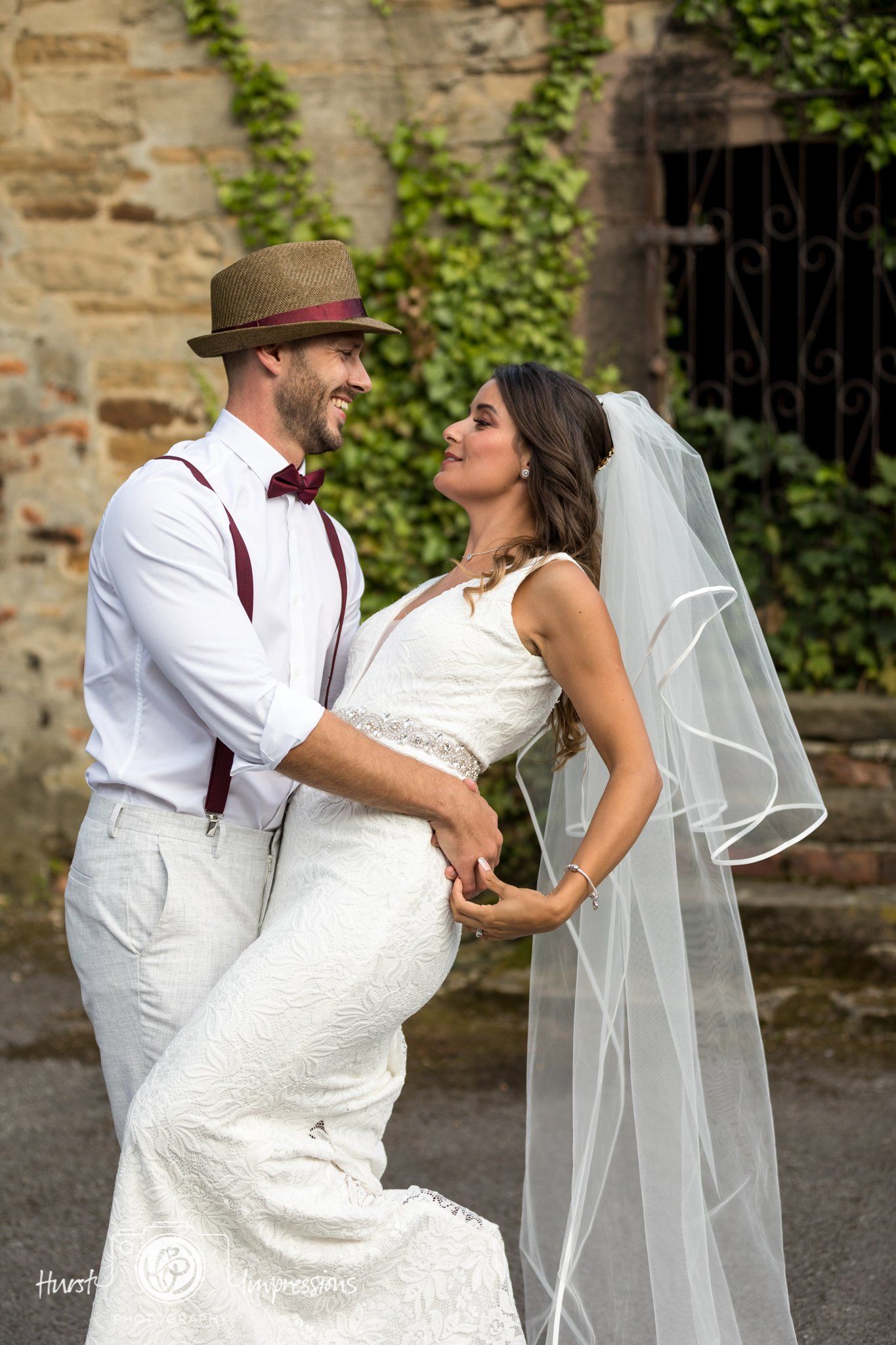 Couple in wedding attire embrace, smiling, outdoors; stone building and greenery in background.