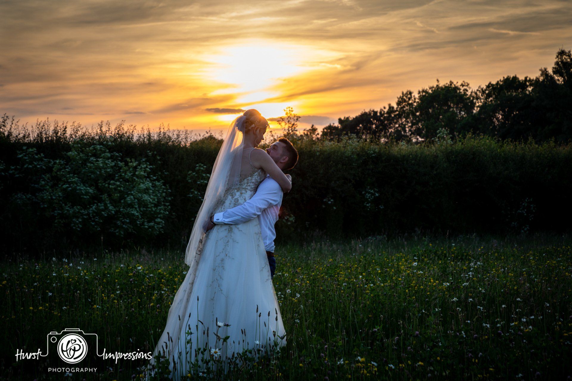 Couple embracing in a field at sunset; woman in wedding dress, man in suit.
