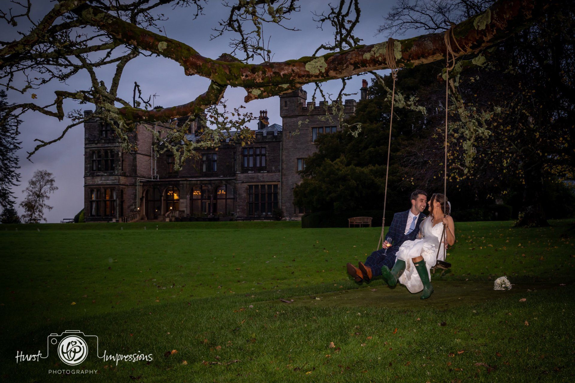 Couple on a swing beneath a tree, in front of a castle at dusk.