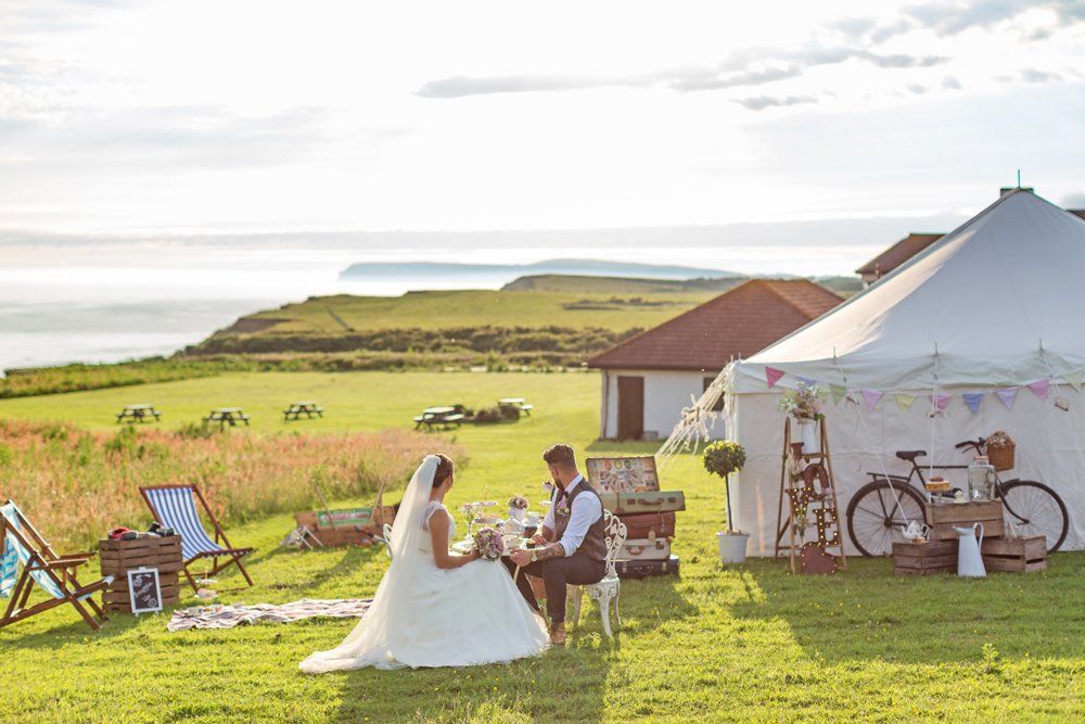 Bride and groom at a table in a field by the ocean. White tent and chairs nearby, sunny.
