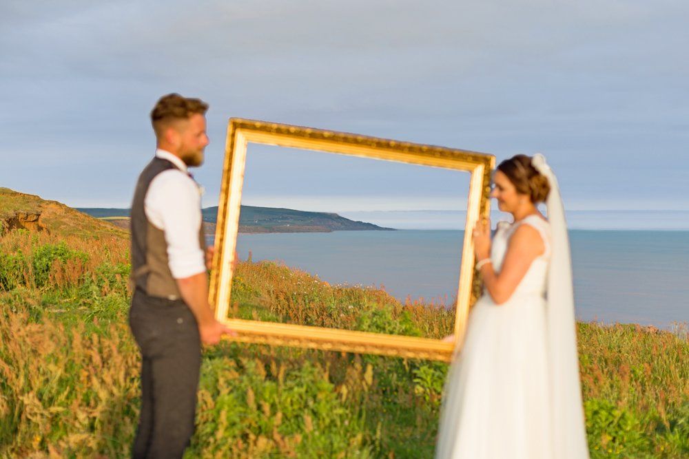 Wedding couple holding a gold picture frame overlooking the ocean.