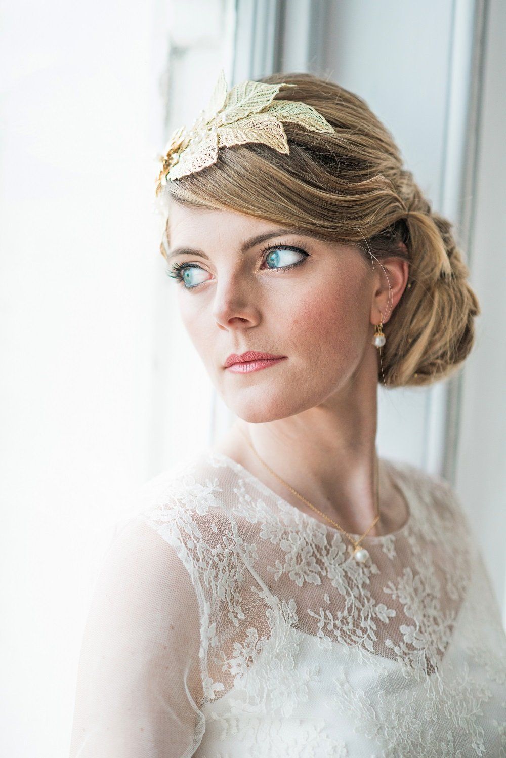 Woman with blonde updo, wearing a lace dress and gold headpiece, looks out a window.