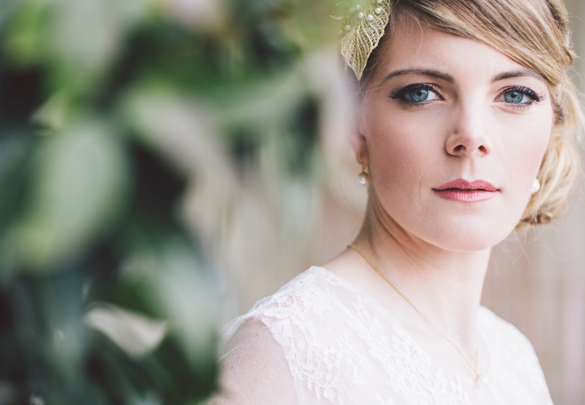 Woman with blonde hair, soft makeup, in a white lace dress; looking at the camera with a serious expression; outdoors.