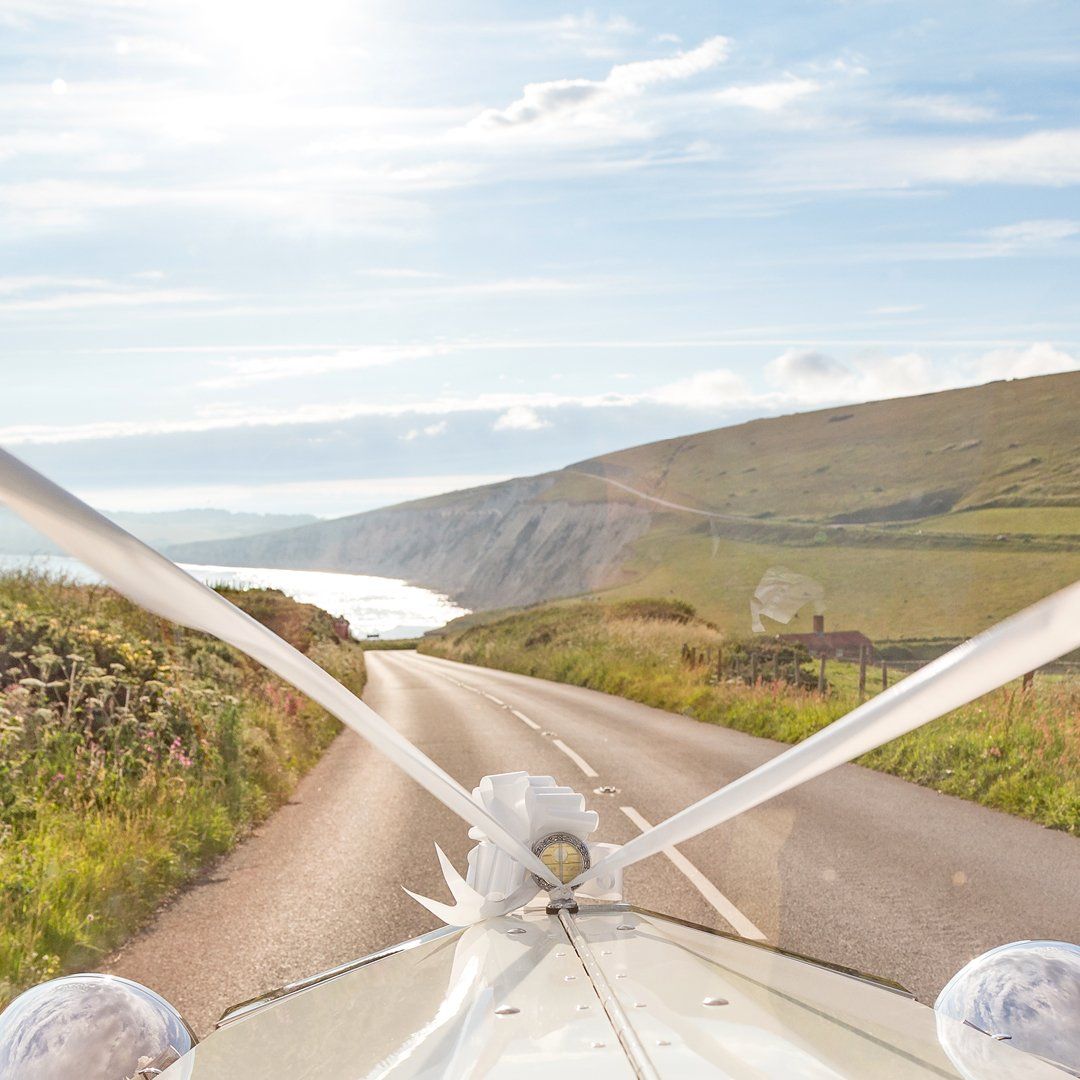 Airplane wing struts frame a coastal road winding along a green hillside under a blue sky.