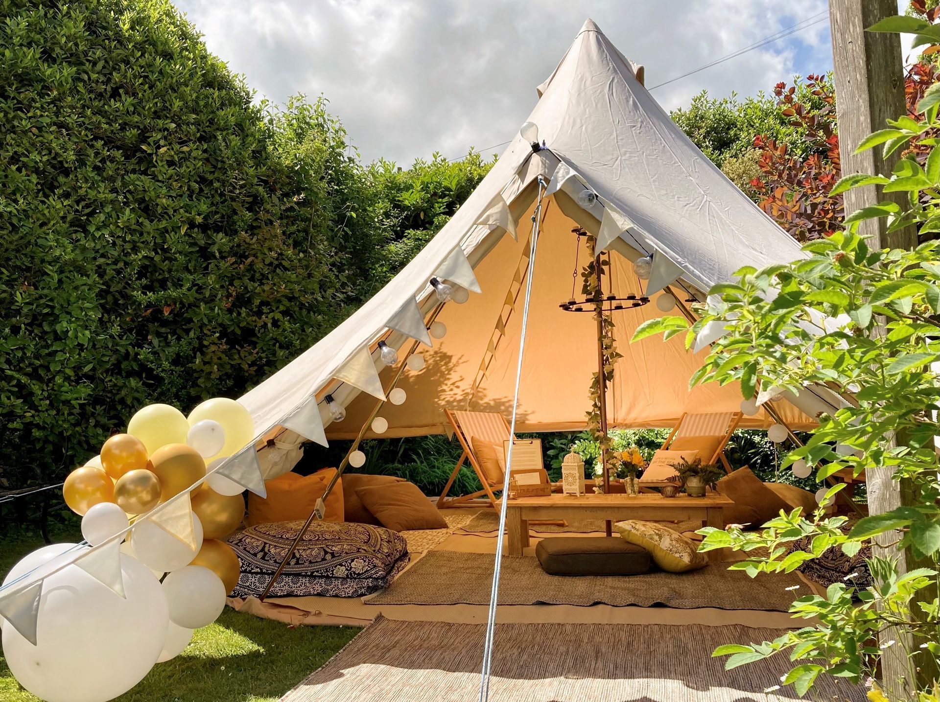 Bell tent with bunting, balloons, and cushions set up in a garden for a gathering.