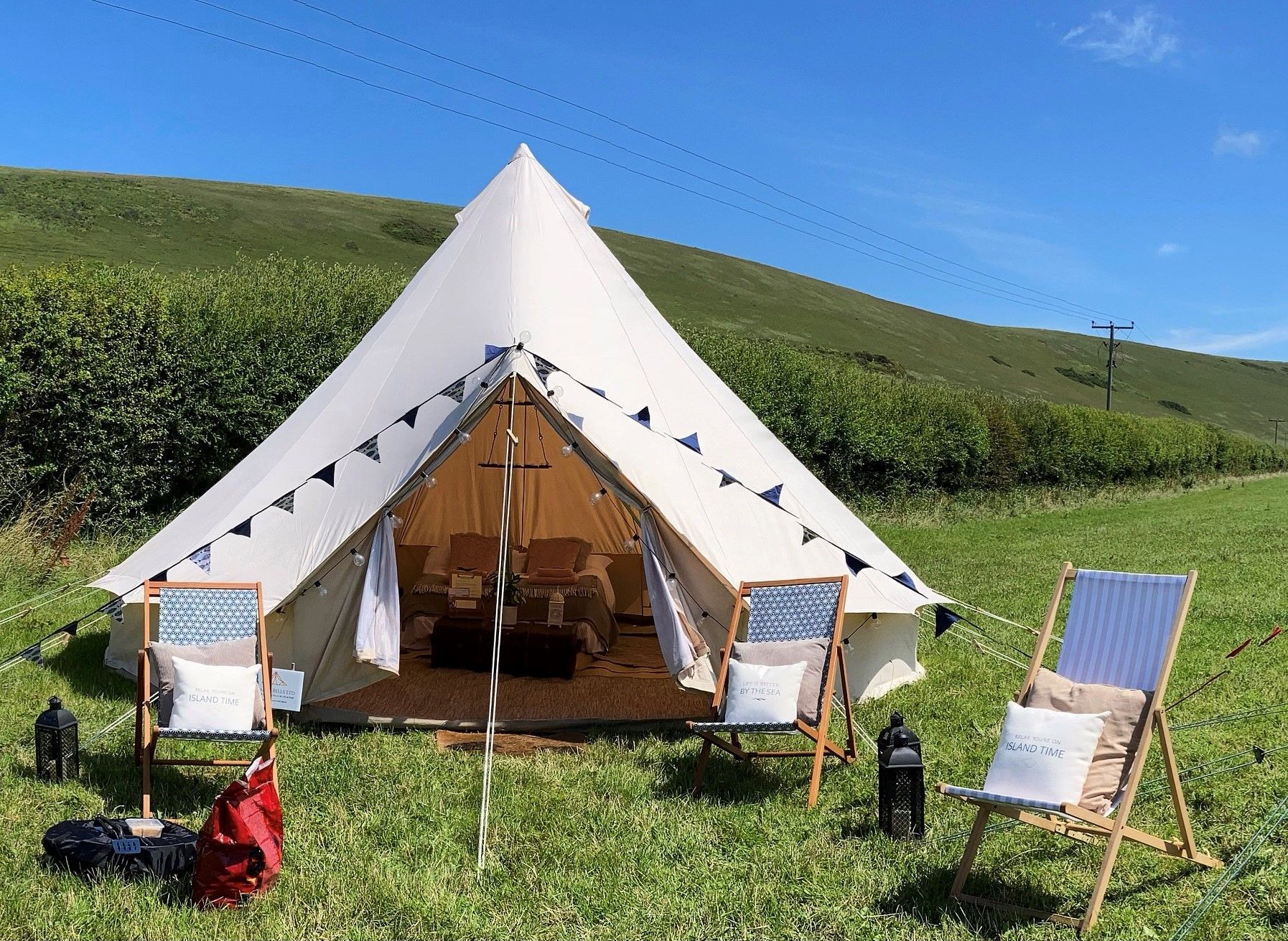 Cream-colored bell tent in a grassy field, decorated with blue and white bunting and chairs, with rolling hills in the background.