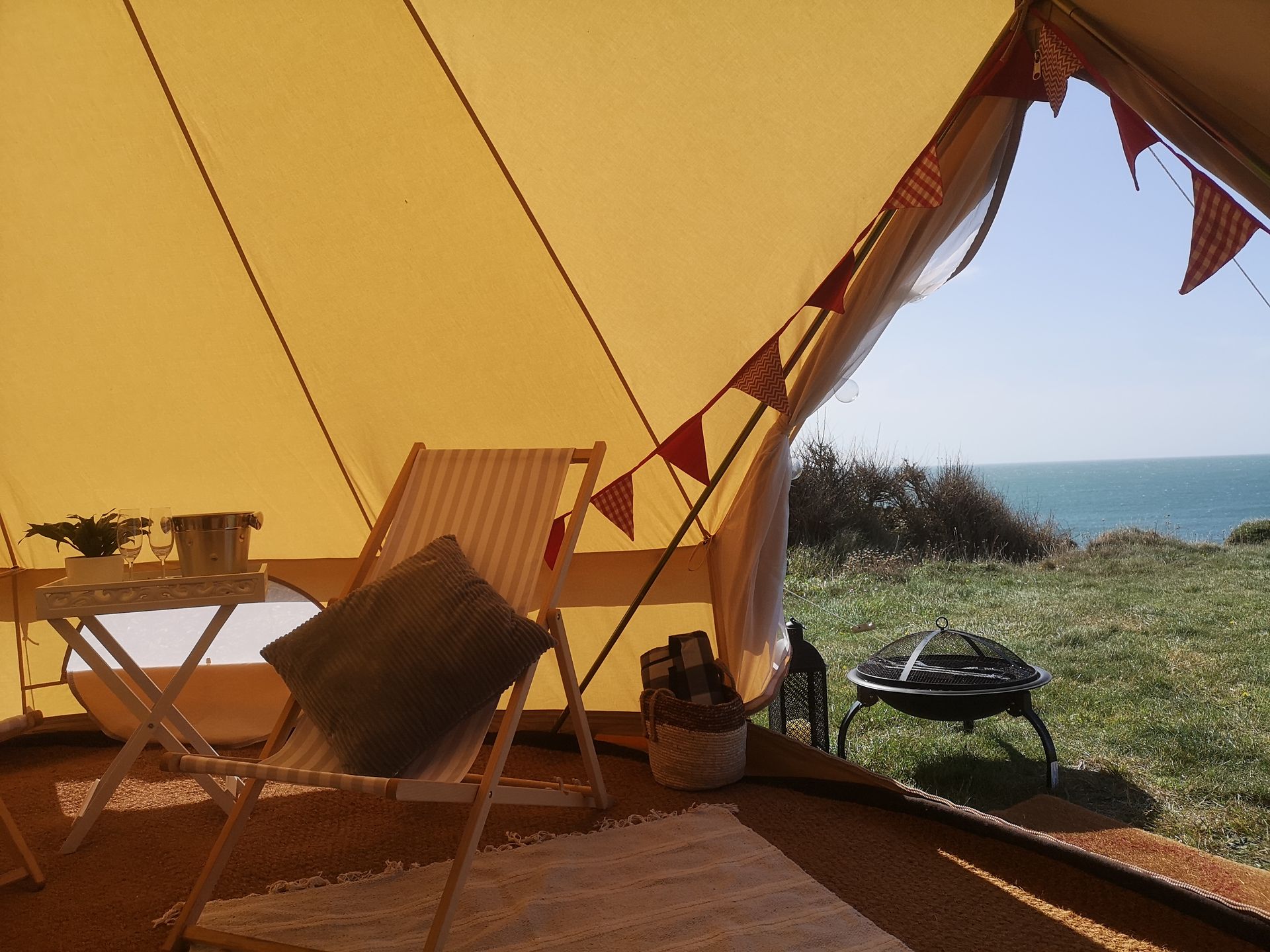 View from inside a tent, looking out at a grassy cliffside and ocean. Includes chairs, a table, and a fire pit.