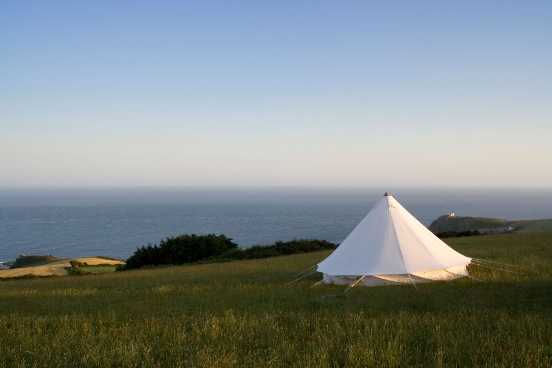 White bell tent on a grassy hillside overlooking the ocean. Blue sky overhead.
