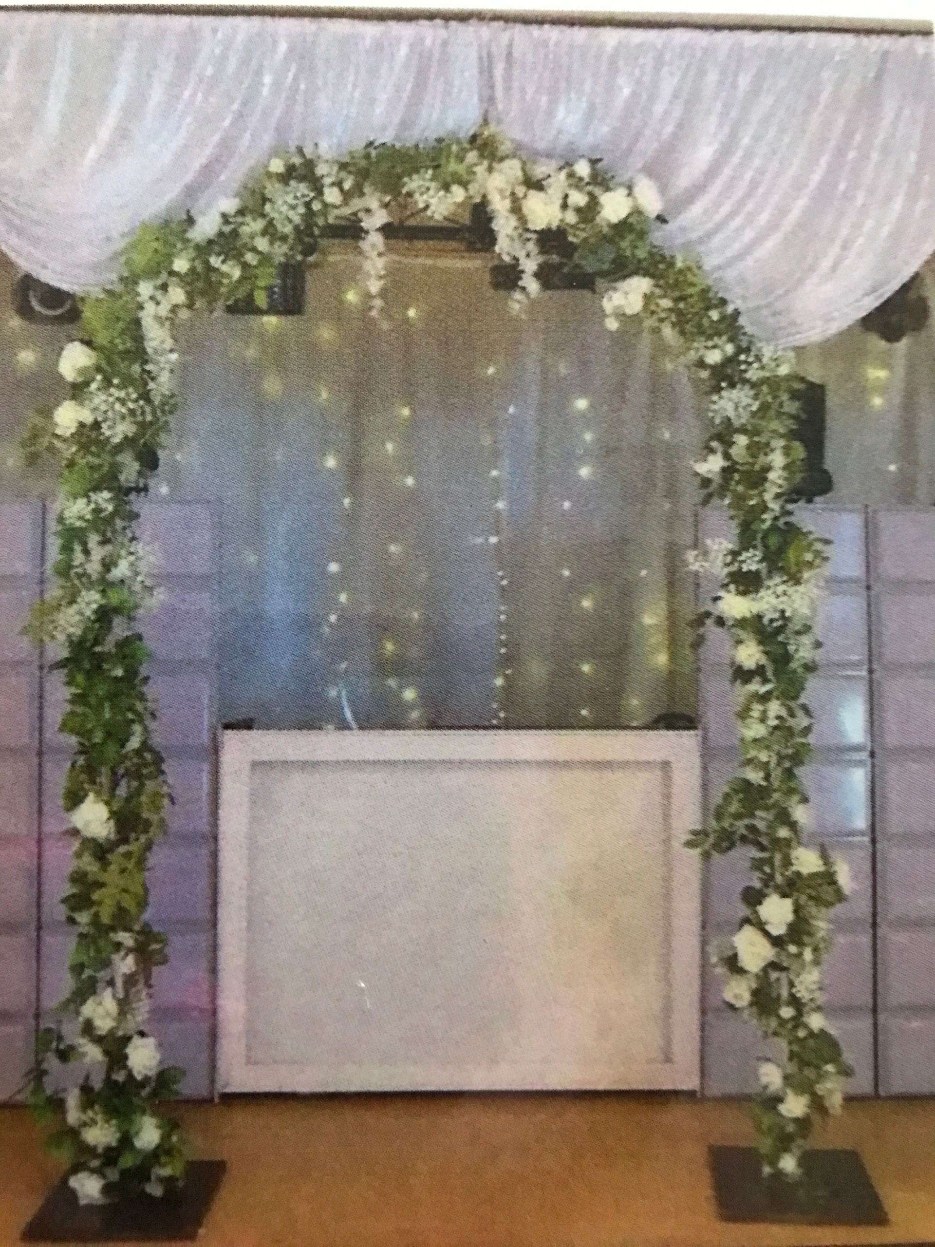 Floral archway with white roses and greenery, backdrop with fairy lights.