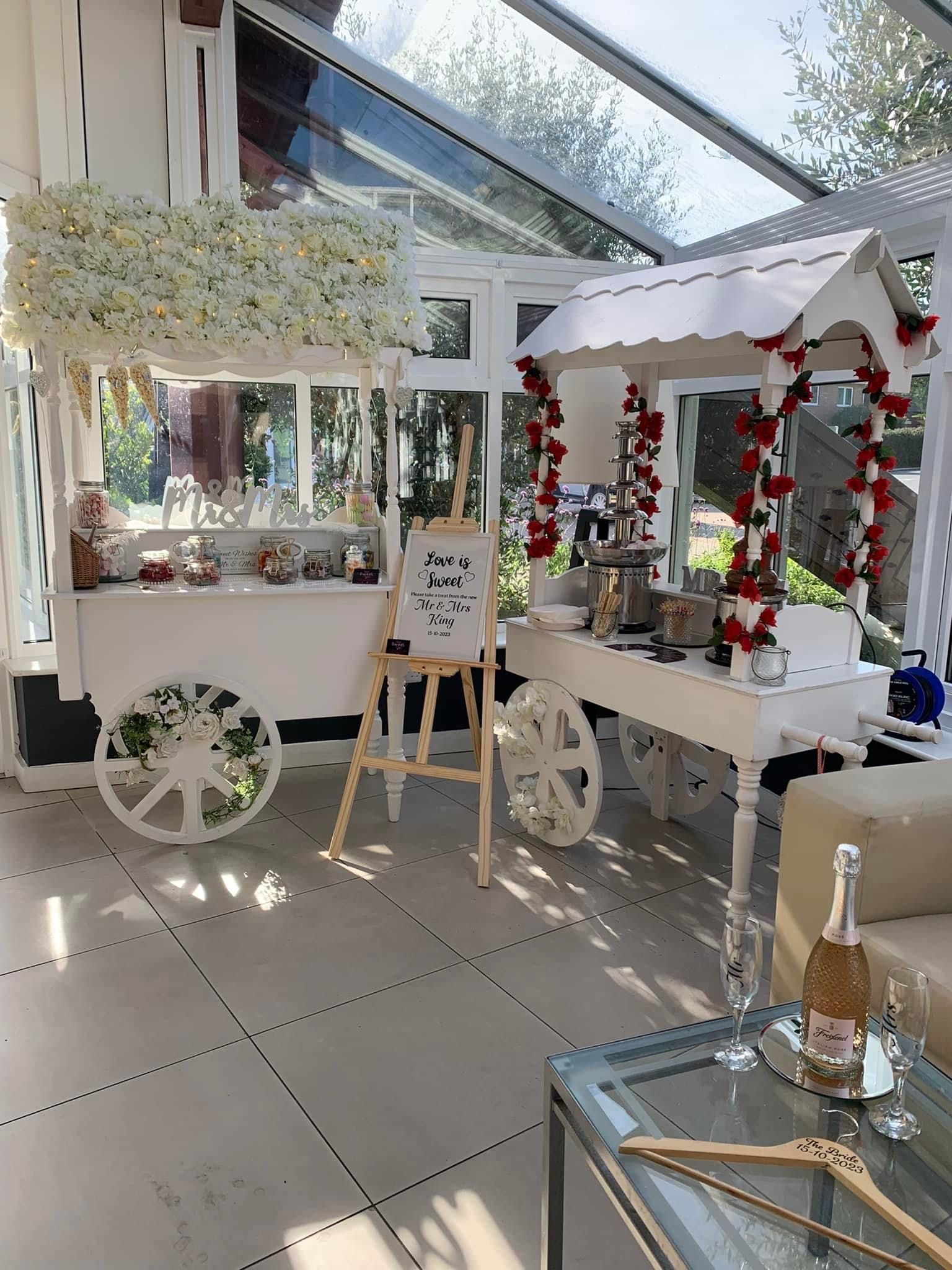 White dessert carts with treats in a sunroom; one has flowers, the other, red roses. A welcome sign stands between them.