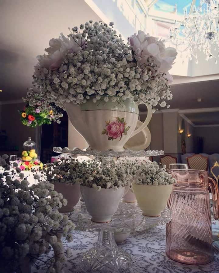 Floral arrangement in teacups and a teapot filled with baby's breath and peonies, on a table set for an event.