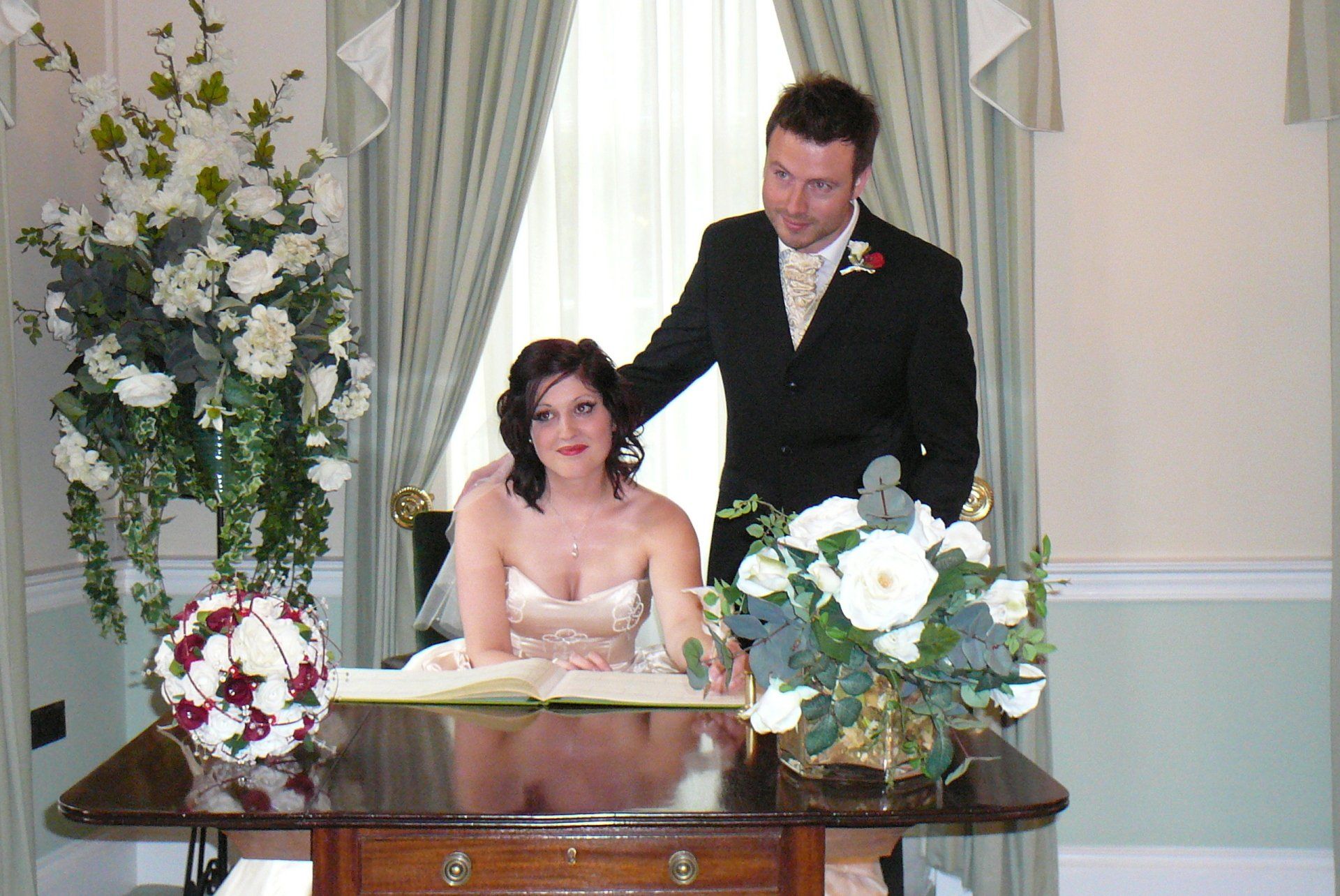 Bride and groom signing marriage certificate at a table in a room with white and green floral decorations.