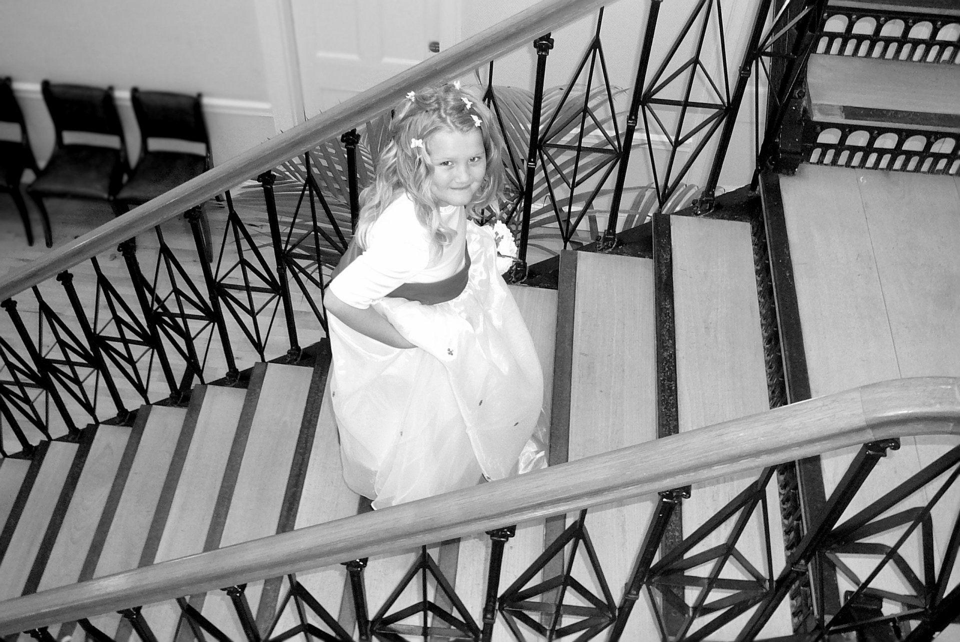 Girl in formal dress on a staircase with ornate railing, looking up. Black and white.