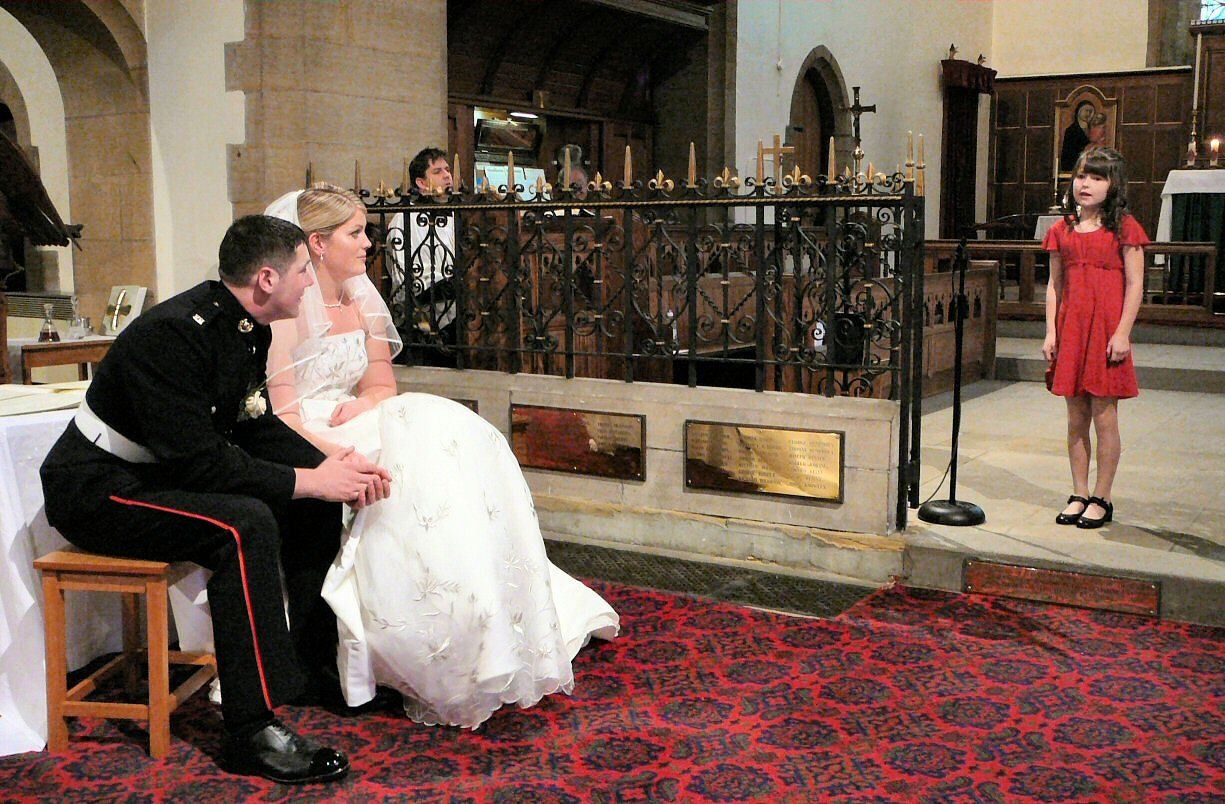 Wedding ceremony: Couple seated as girl sings in church, red carpet, dark uniforms.