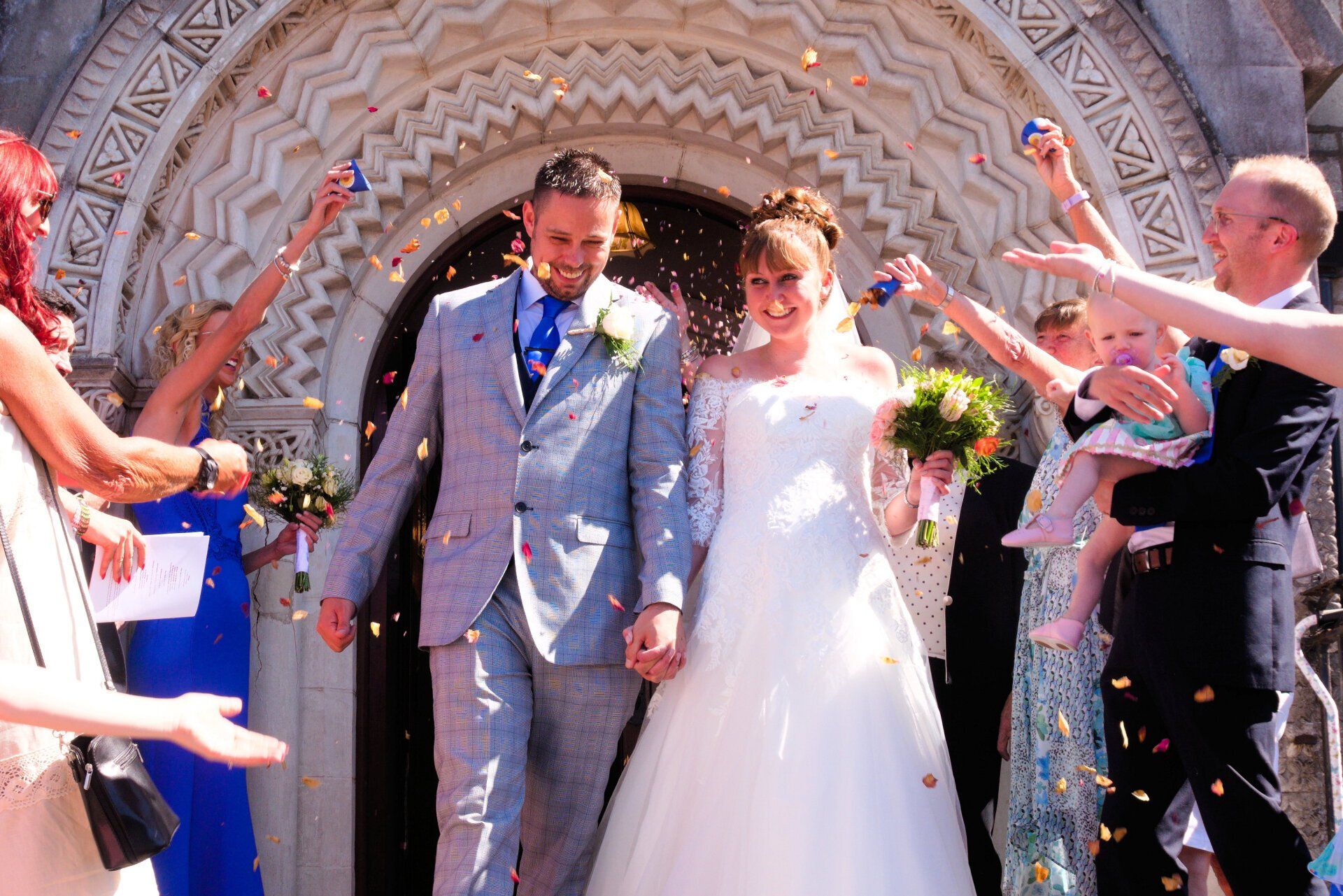 Newlyweds exiting a church, showered with confetti. Groom in a gray suit, bride in a white gown. People celebrate.