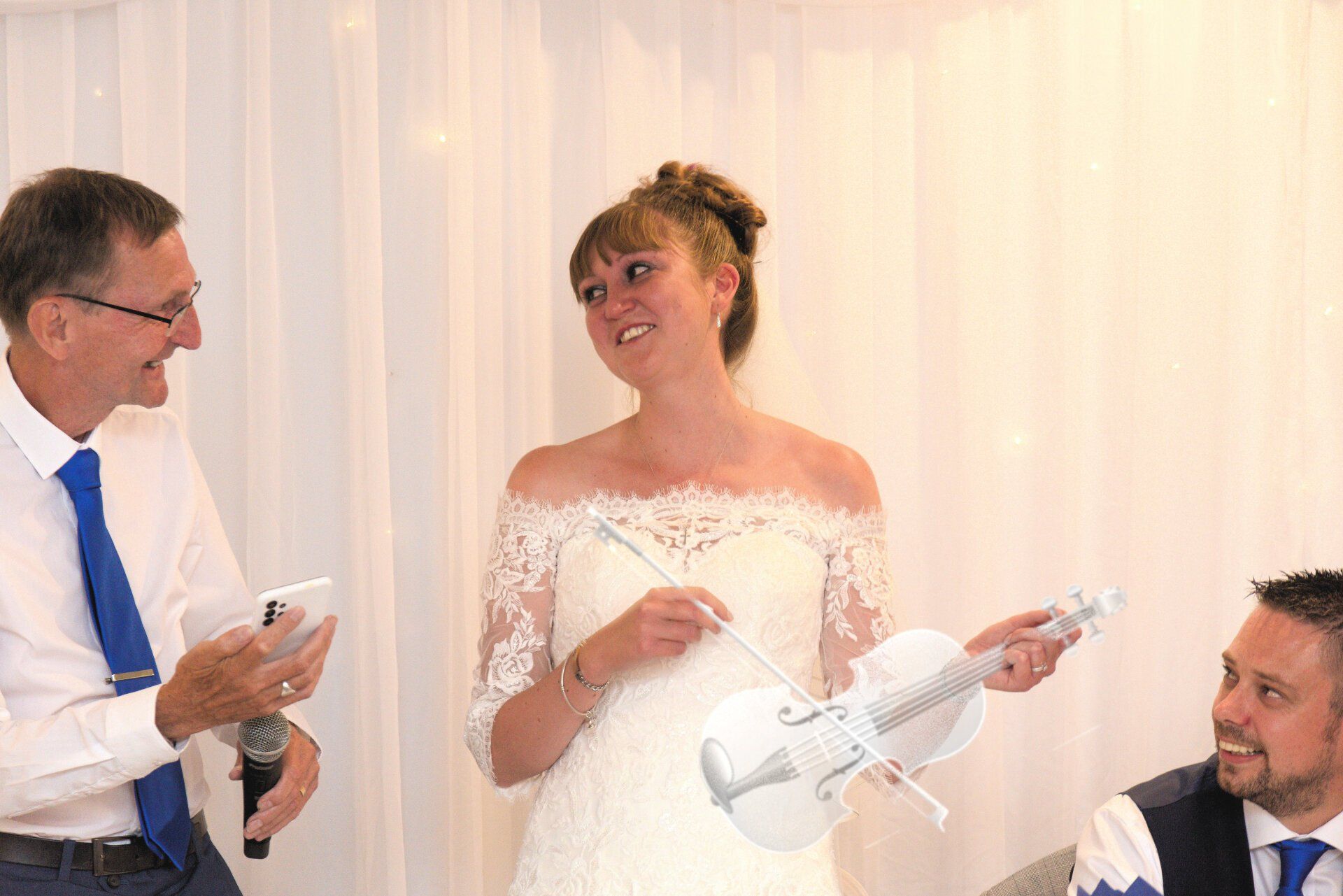 Bride with toy violin smiles, as man on left holds microphone, and another man looks on.