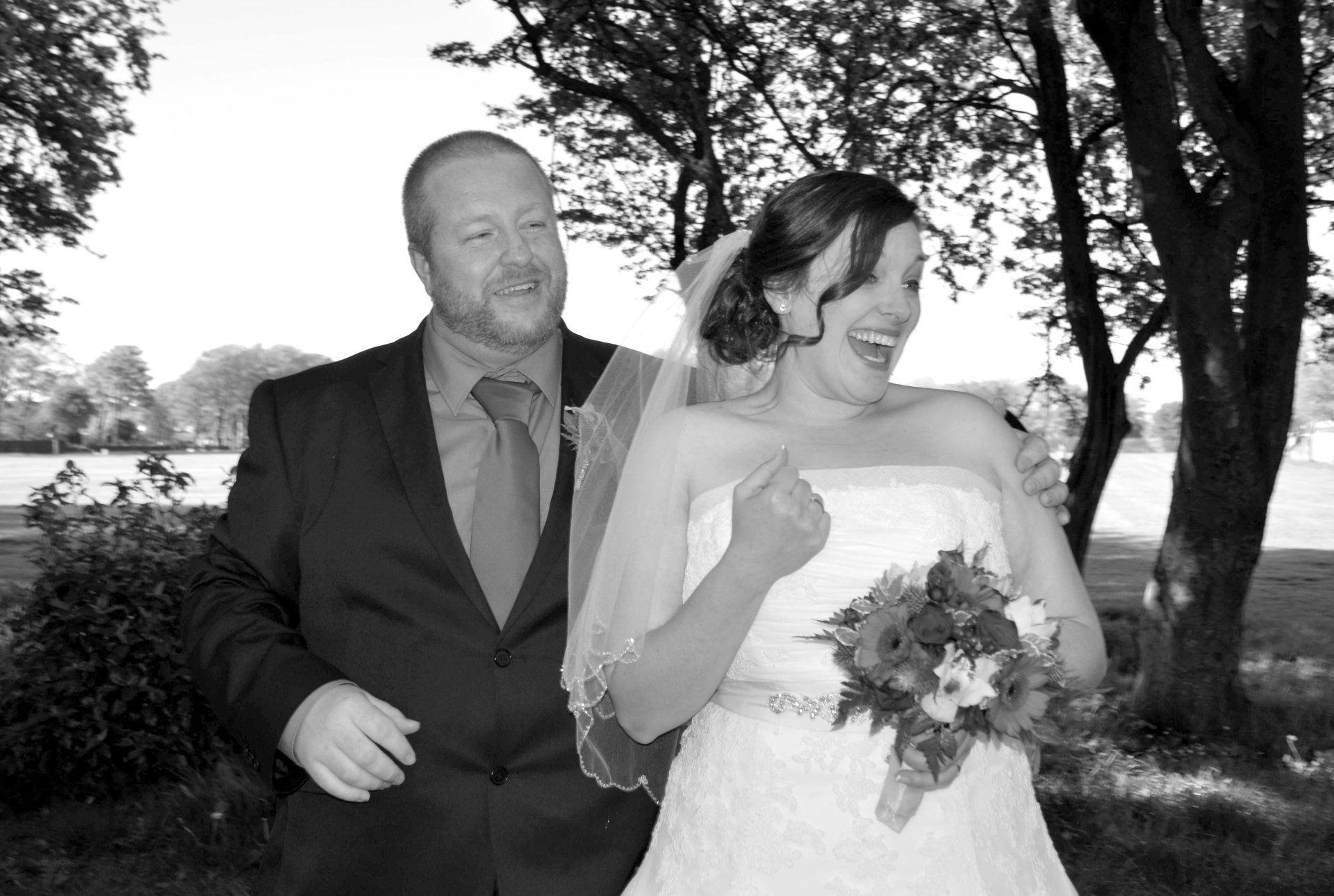 Groom smiles, arm around laughing bride in a strapless wedding dress, holding flowers, trees and field in background.