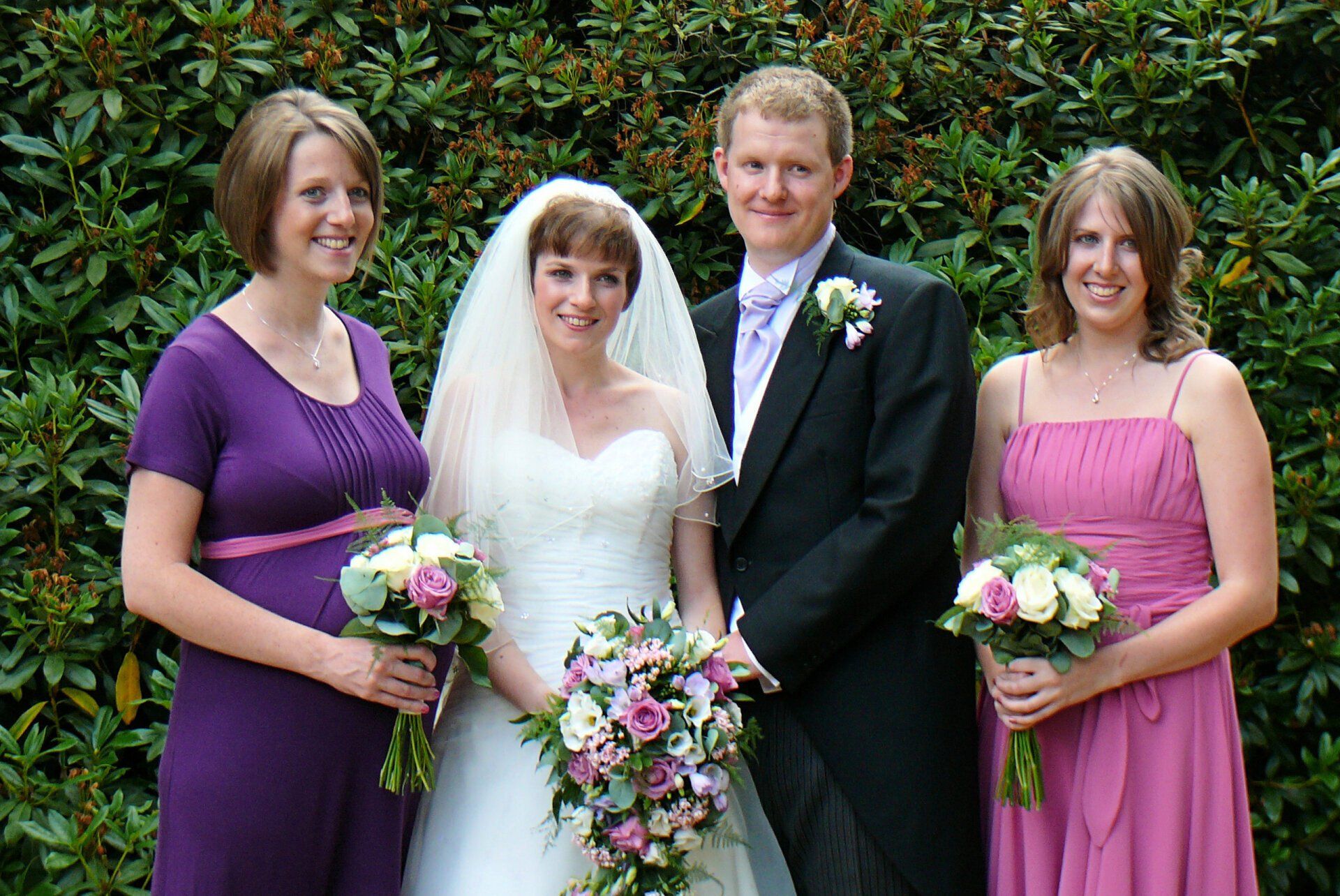 Wedding party: Bride and groom with two bridesmaids, posing outdoors in front of green foliage.