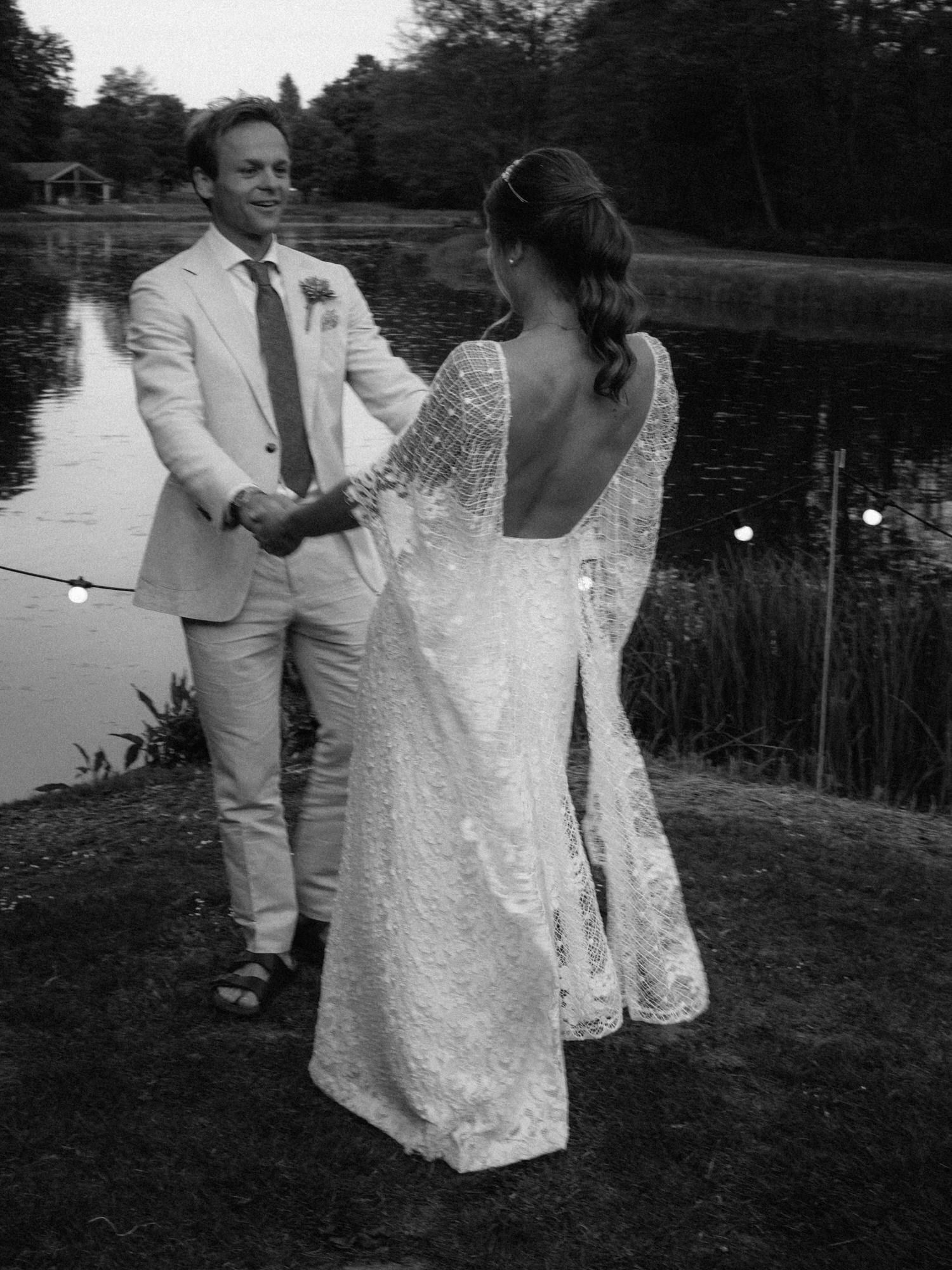 Couple in wedding attire holding hands, by a lake. Man in a light suit, woman in a lace gown.