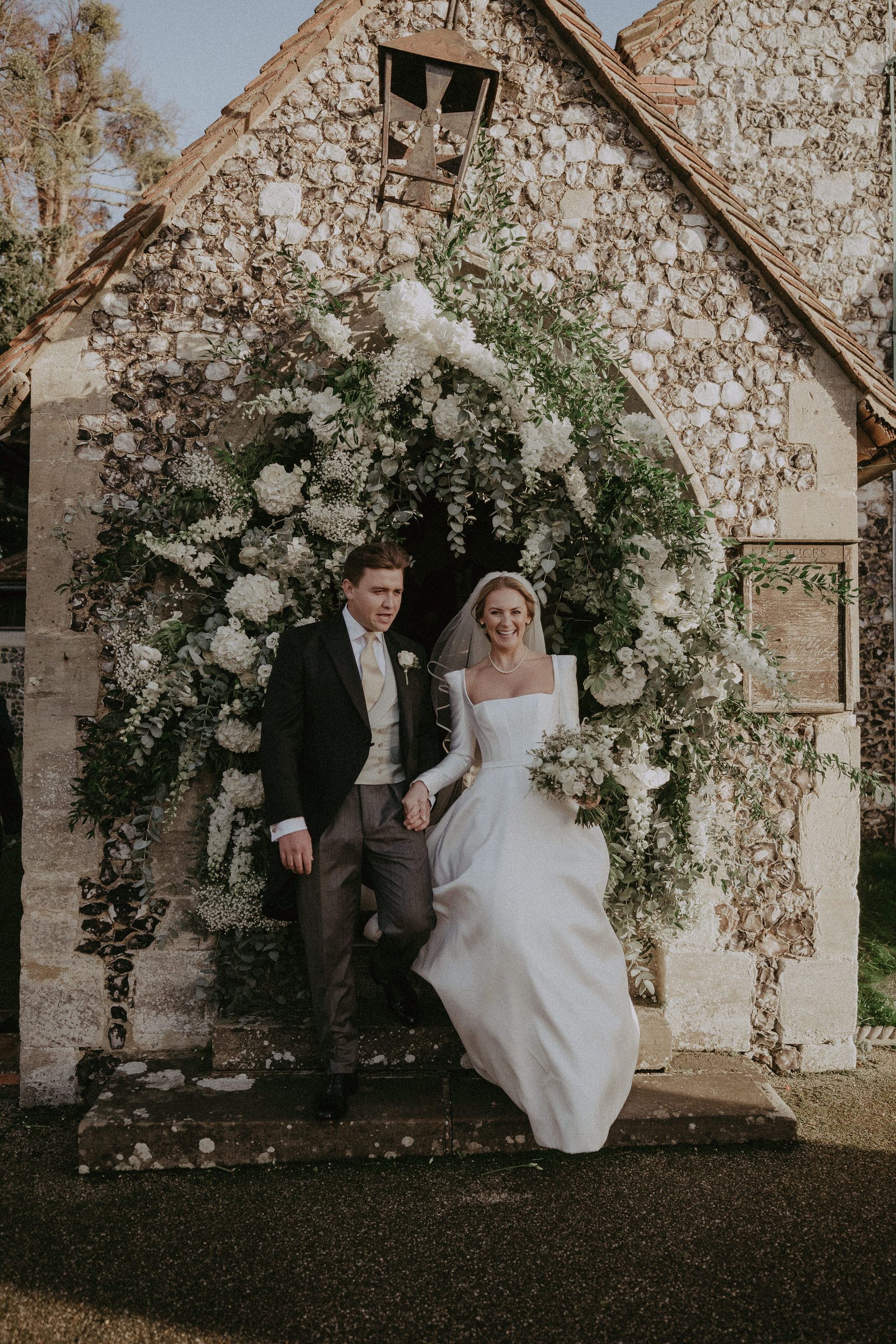 Bride and groom exiting a stone church under a floral archway.