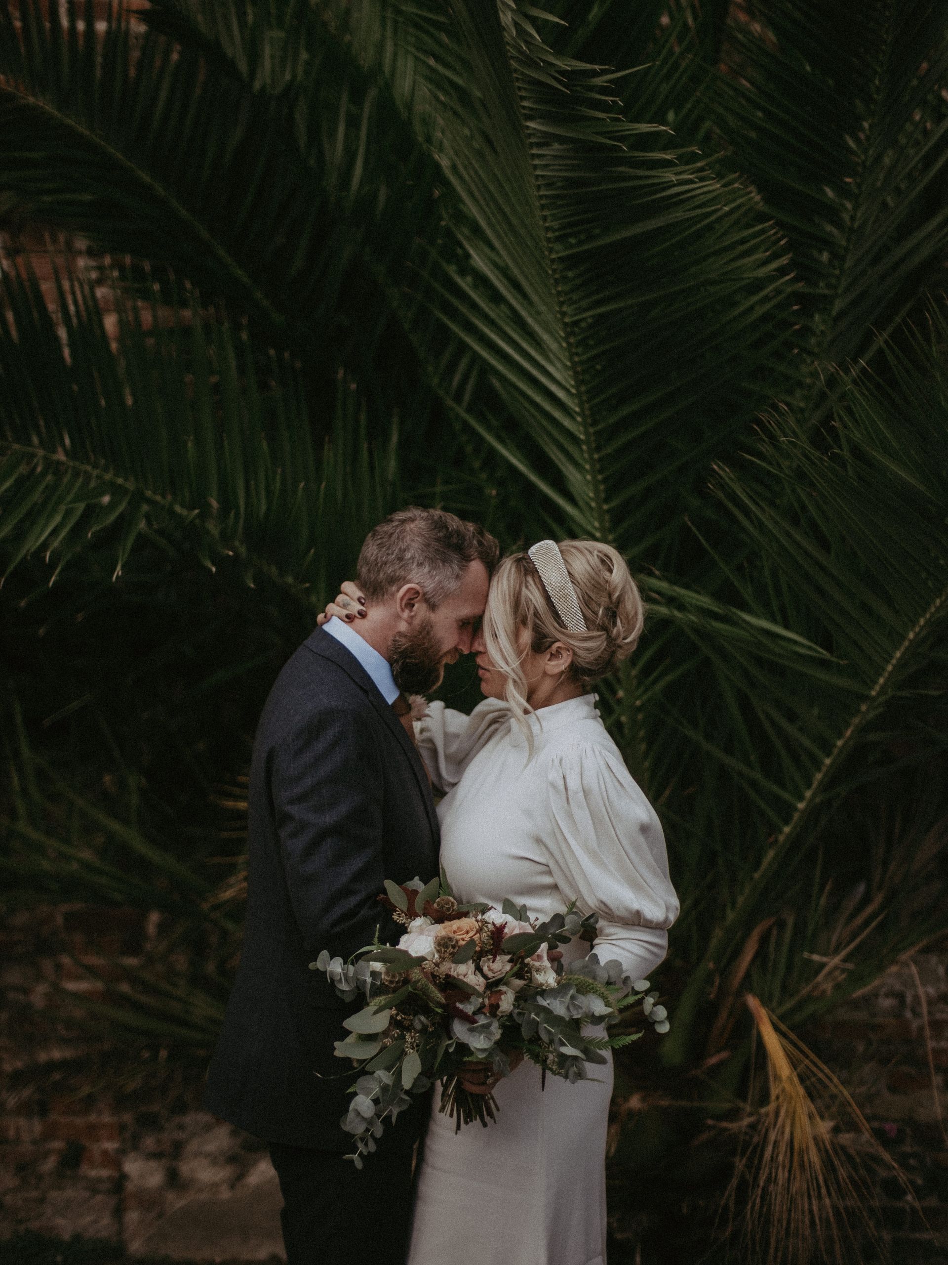 Couple embraces, bride in white dress, groom in suit, palm leaves background, holding bouquet.