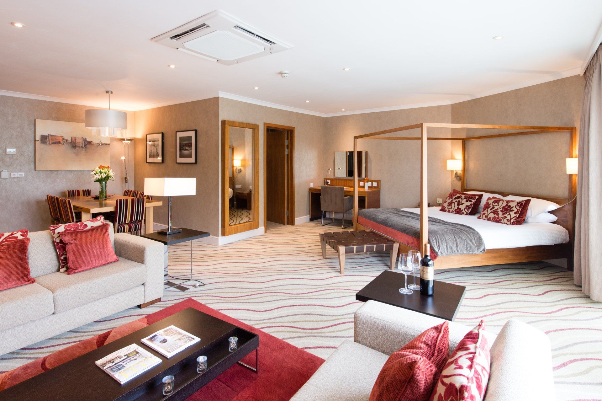 Luxurious hotel suite with a canopy bed, dining area, and seating, featuring neutral tones with pops of red.