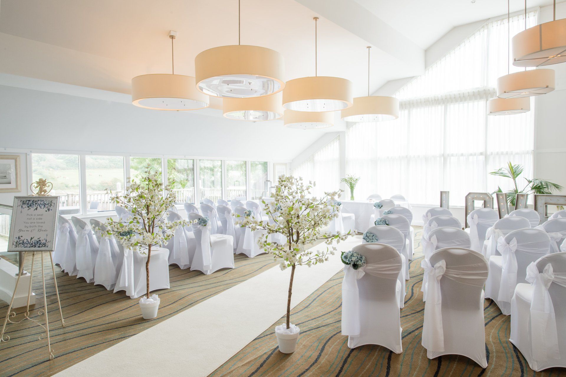 Wedding ceremony room with white chairs, aisle, and large pendant lights.