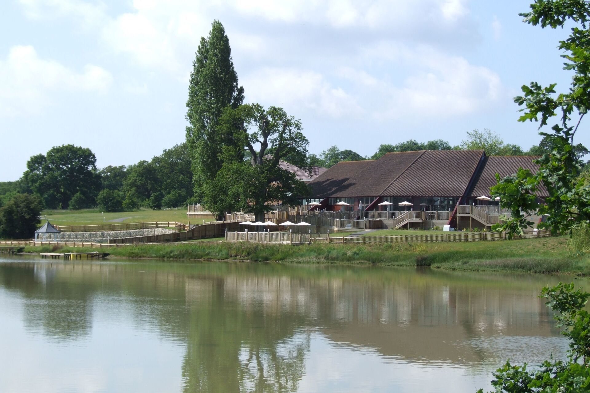 Lakeside building with brown roof, trees, and wooden railings reflected in calm water under a blue sky.