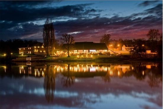Building illuminated at night, reflected in calm water, against a dusky sky with streaks of color.