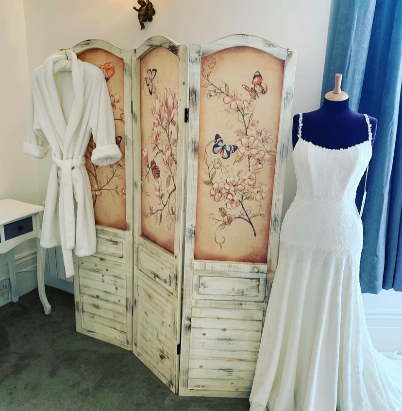 A white wedding dress on a mannequin, a floral screen, and a bathrobe in a room with blue curtains.