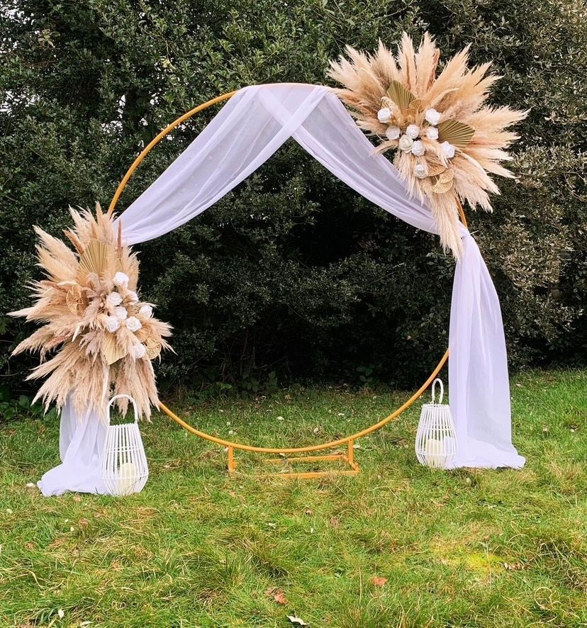 Gold circular wedding arch with pampas grass, white fabric, and lanterns on a grassy lawn.