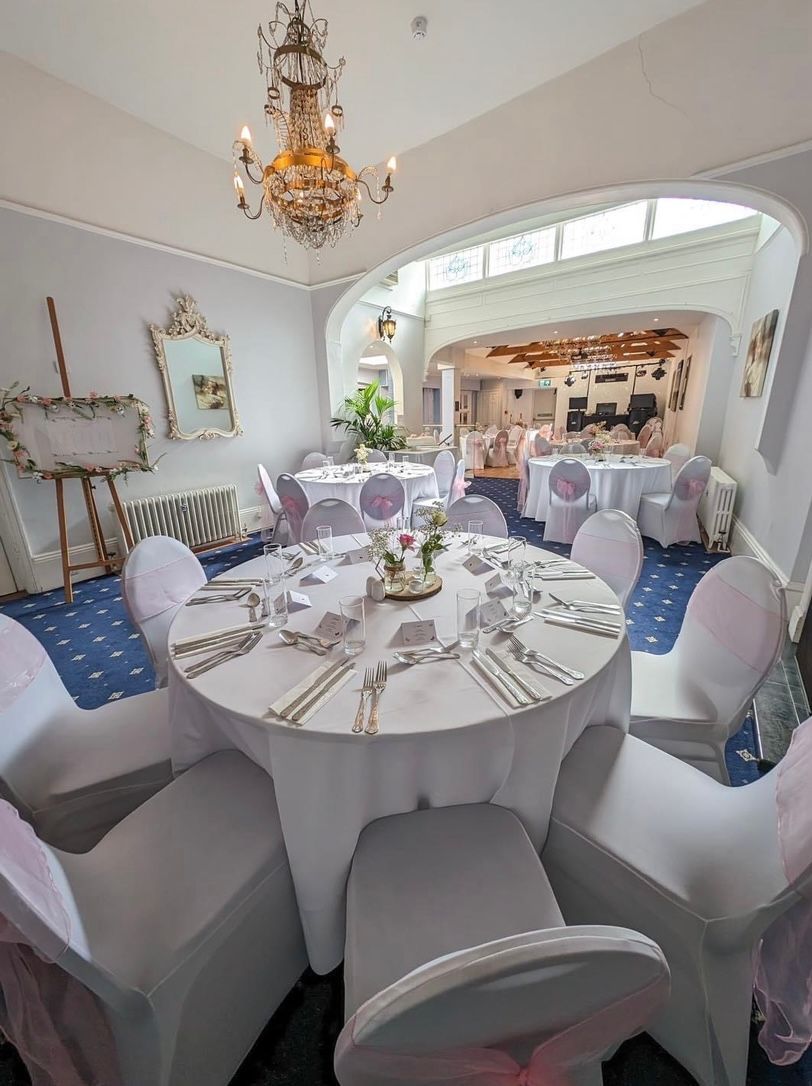Formal dining room with round tables set for a celebration. Chairs have white covers and pink accents; ornate chandelier hangs.