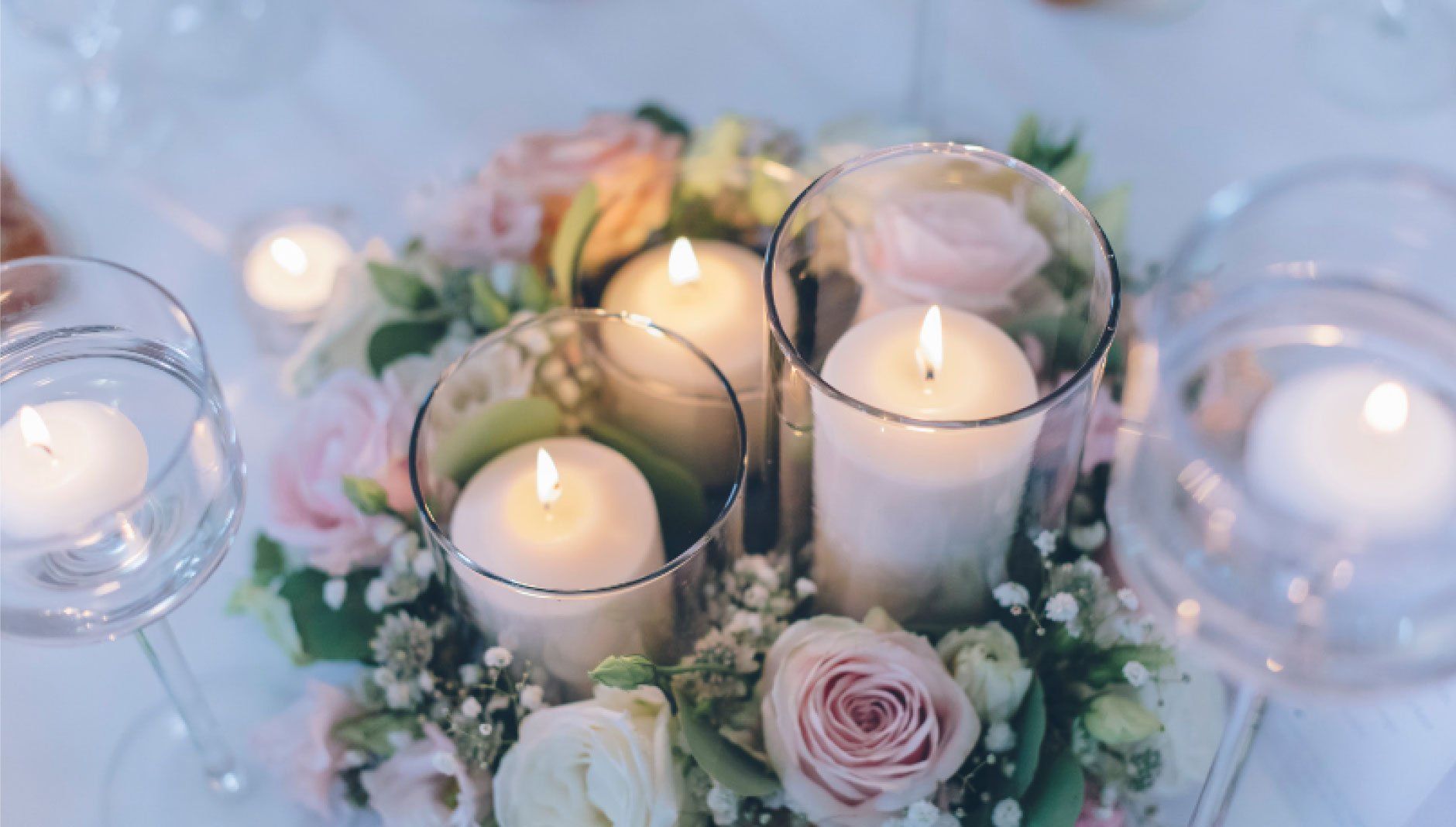 Centerpiece with lit candles, blush roses, greenery in glass containers on a table.