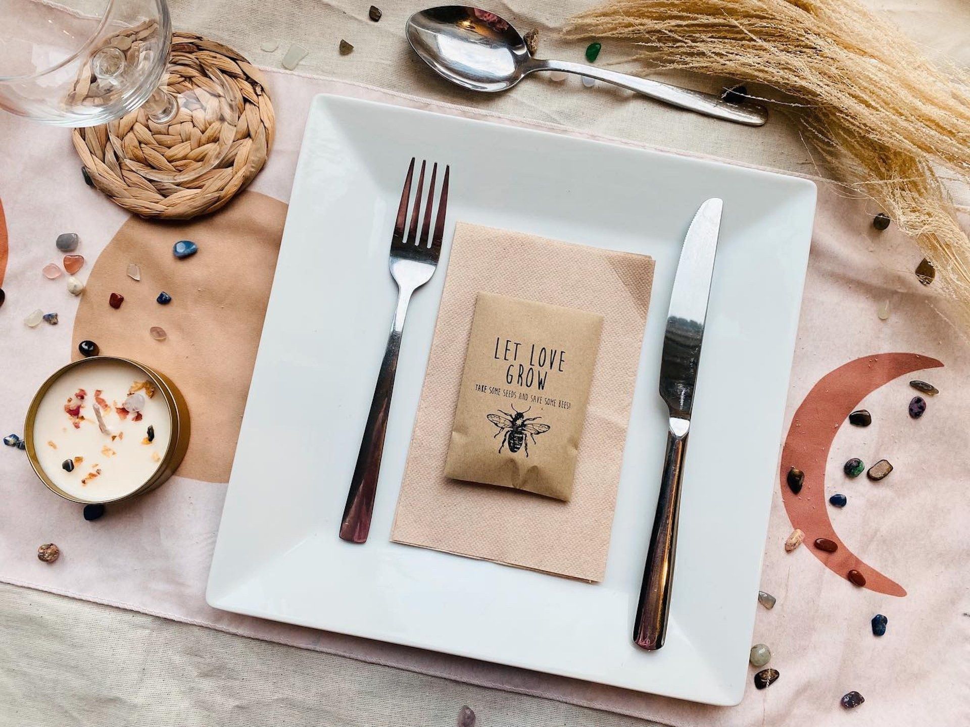 A place setting with a square white plate, silverware, napkin, and a small packet on a table decorated with crystals and a candle.