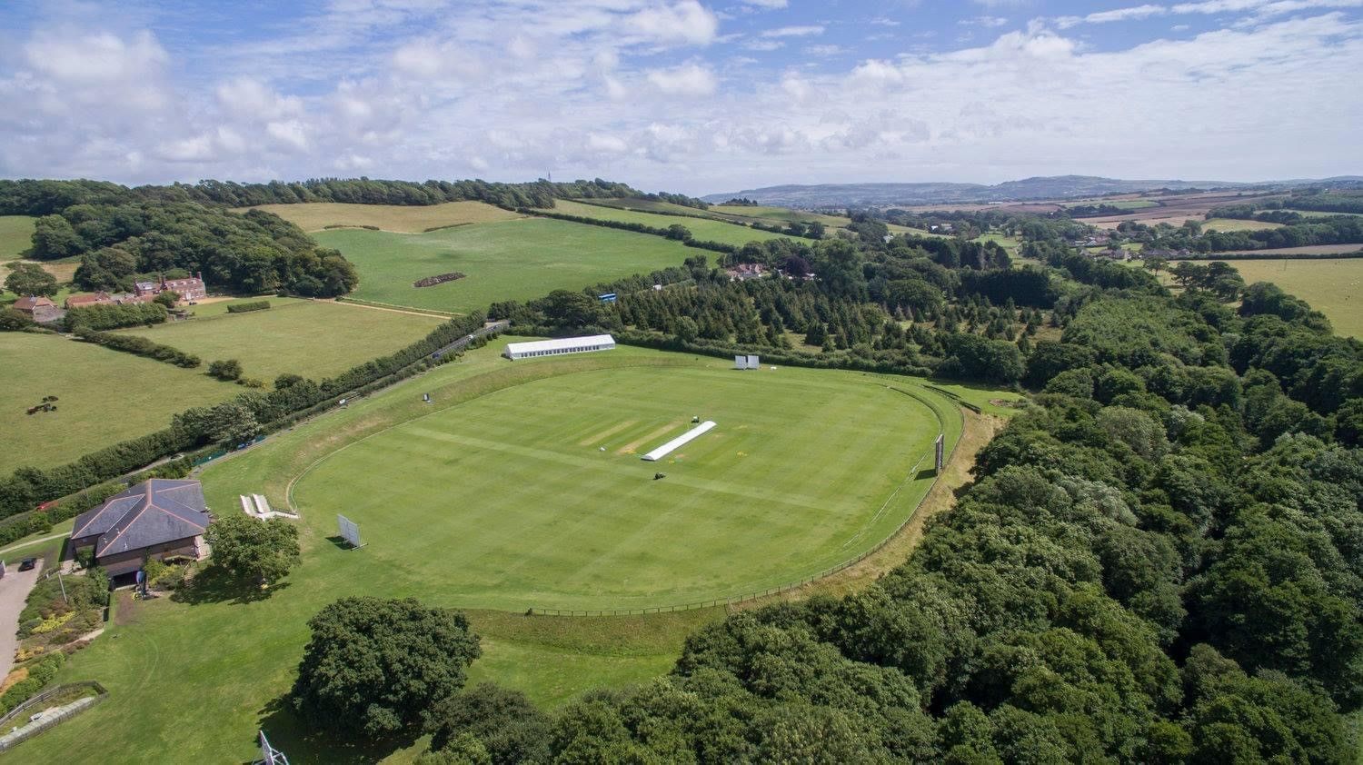 Aerial view of a cricket field surrounded by green fields, trees, and buildings under a blue sky.