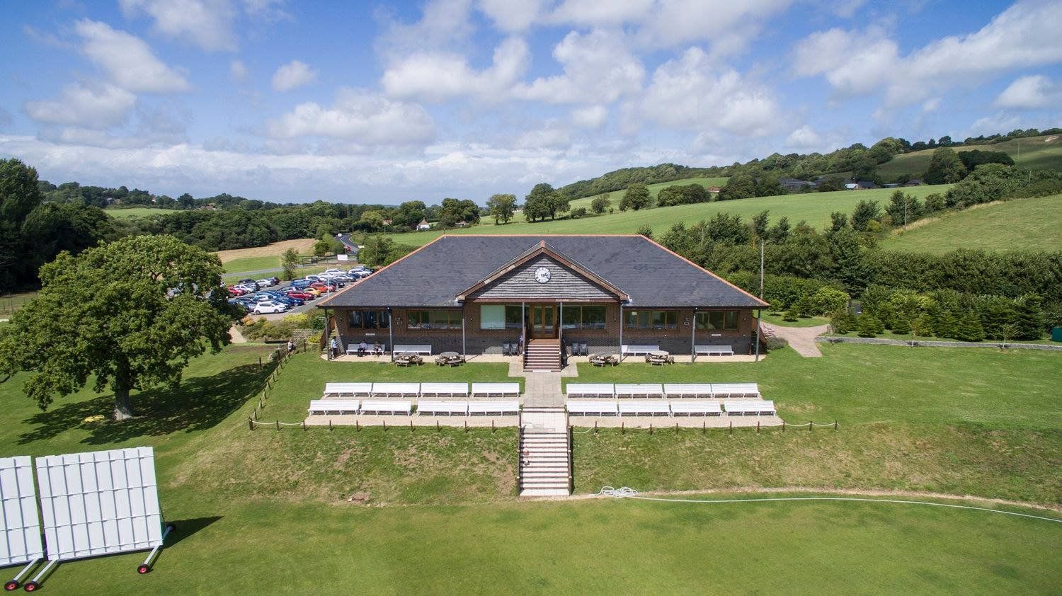 Cricket pavilion on a grassy hill with seating, surrounded by fields and trees under a blue sky.