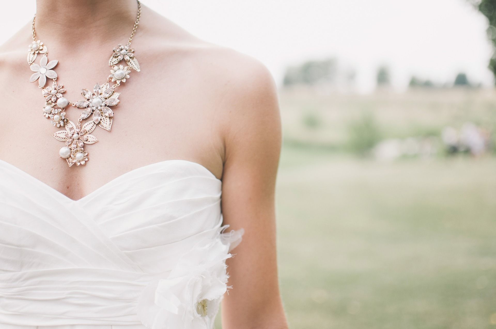 Bride in strapless white dress and ornate necklace; outdoor setting.