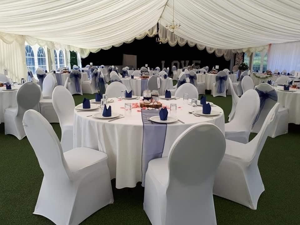 Inside a wedding reception tent, round tables set with white linens, blue accents, white chair covers.