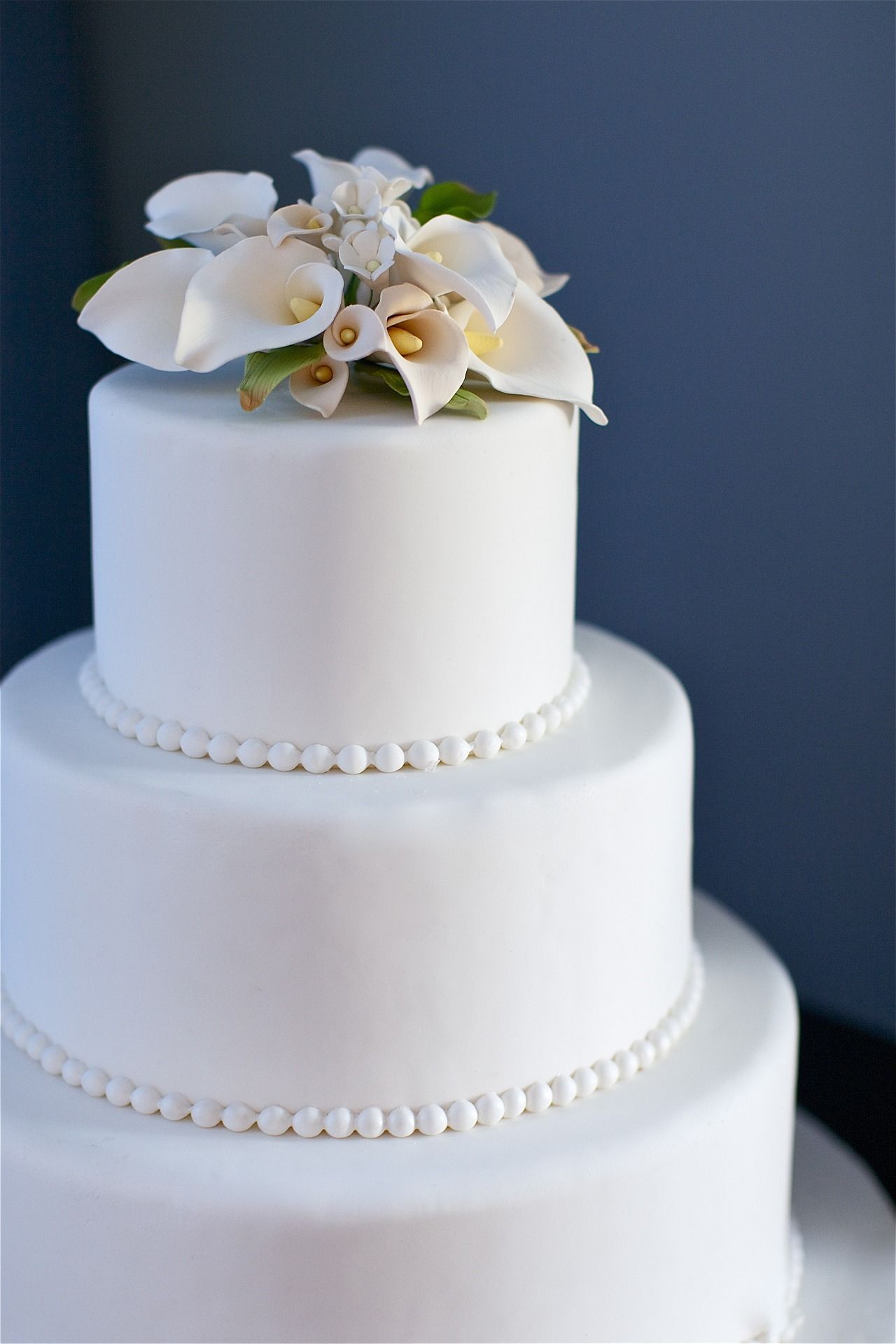 Three-tiered white wedding cake with white calla lily flower arrangement on top and pearl bead borders.